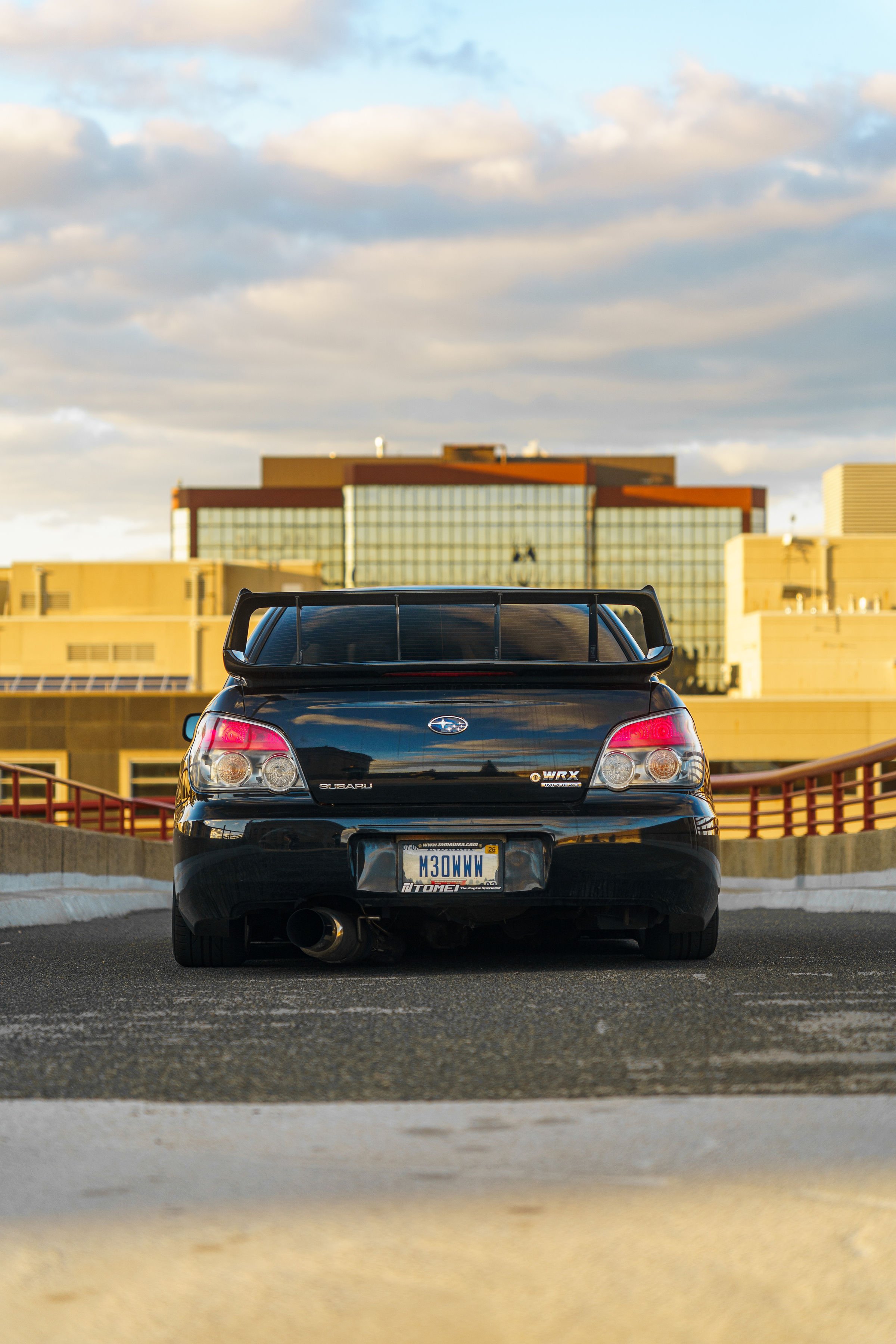 Black Subaru WRX sports car with a rear spoiler, parked outdoors on a parking lot during daytime with a cityscape and cloudy sky in the background.