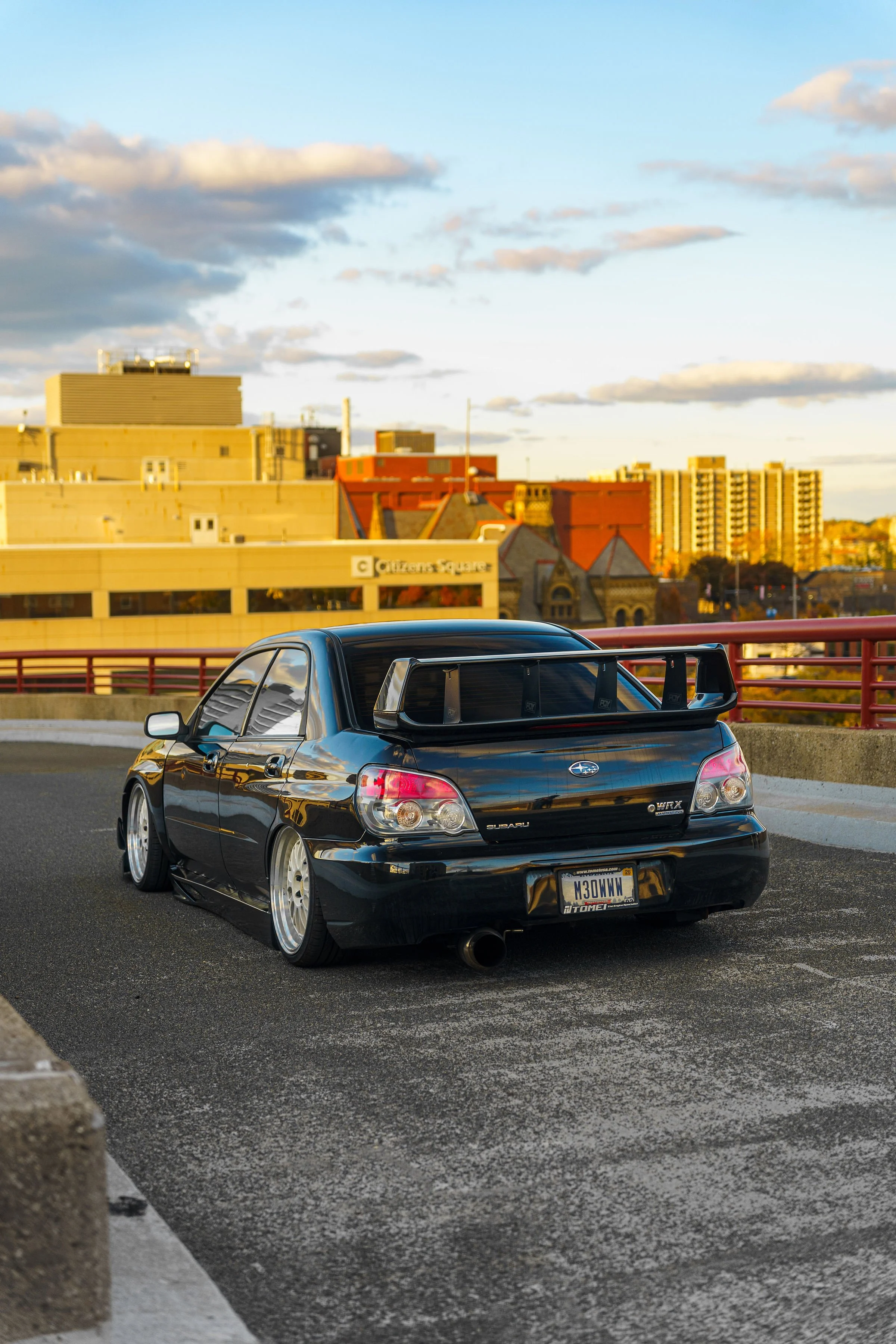A black Subaru WRX car parked on a rooftop parking lot with a cityscape background showing buildings and a blue sky with clouds.