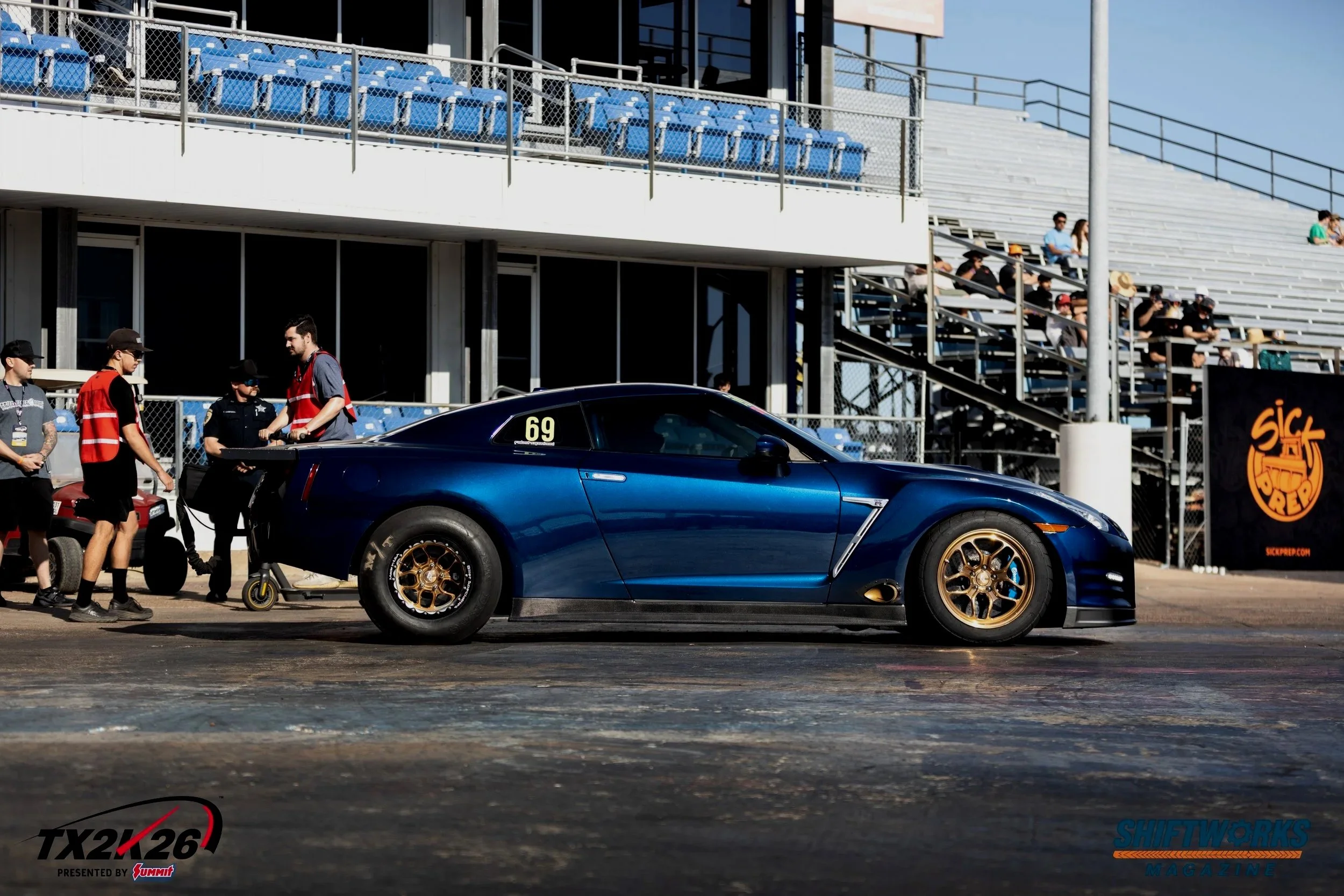 A blue sports car on a racing track with people around it, including crew members and spectators. In the background, there are stadium seating and spectators watching the event.