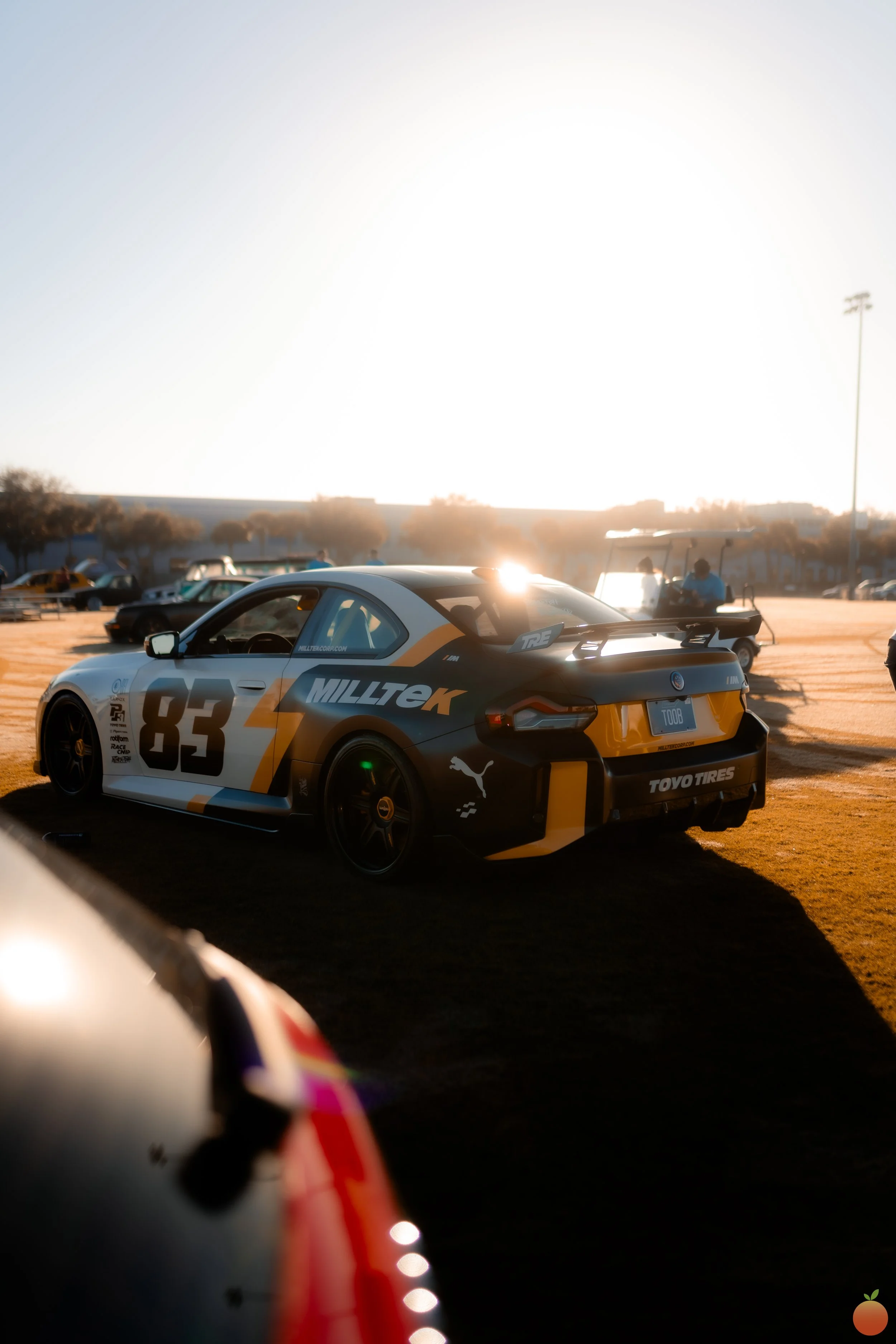 A race car with a yellow, black, and white design, the number 83, and sponsor logos, parked on a dirt lot during sunset with other vehicles and people in the background.