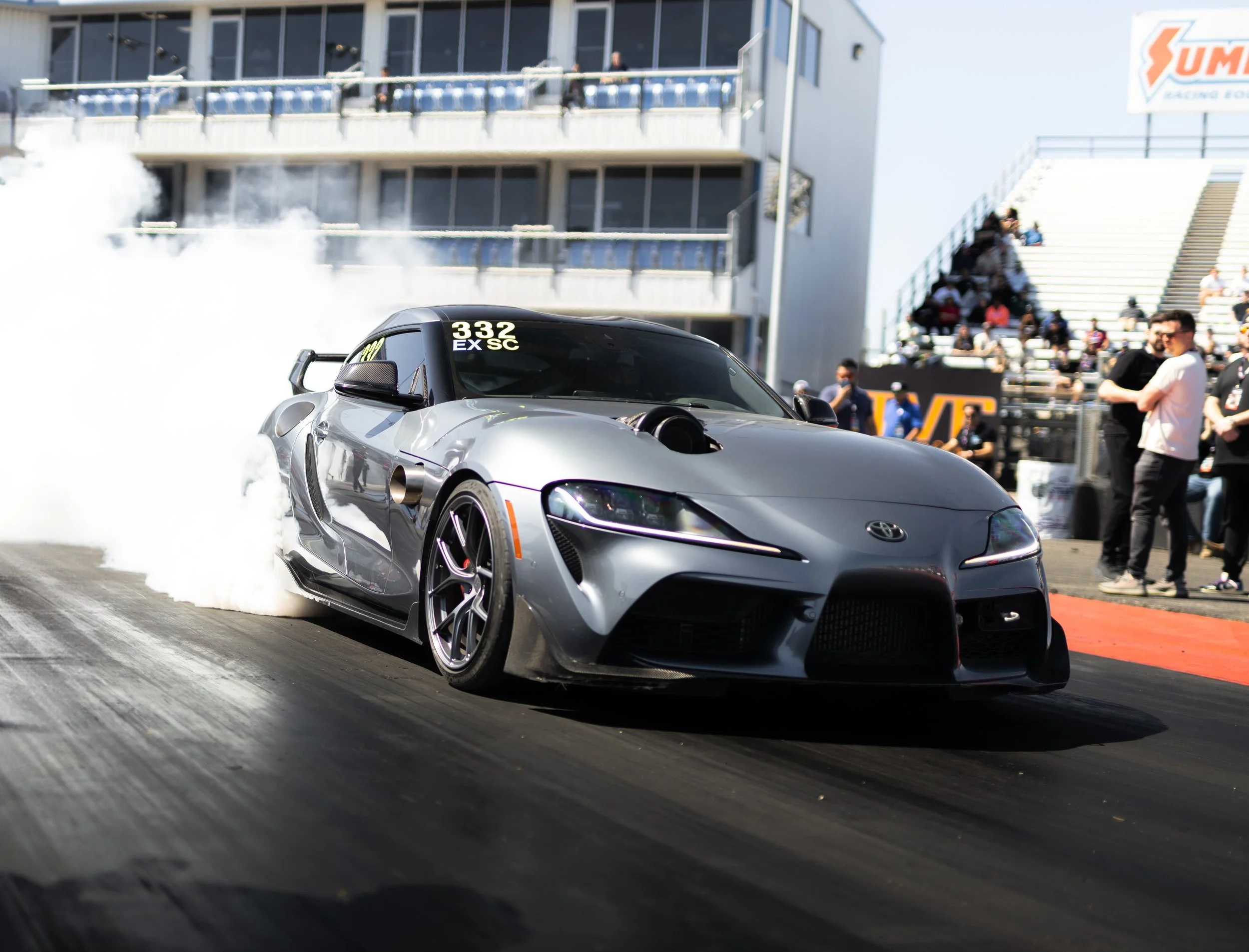A silver sports car performing a burnout at a racing event, with smoke billowing from the rear tires and a crowd of spectators watching nearby.