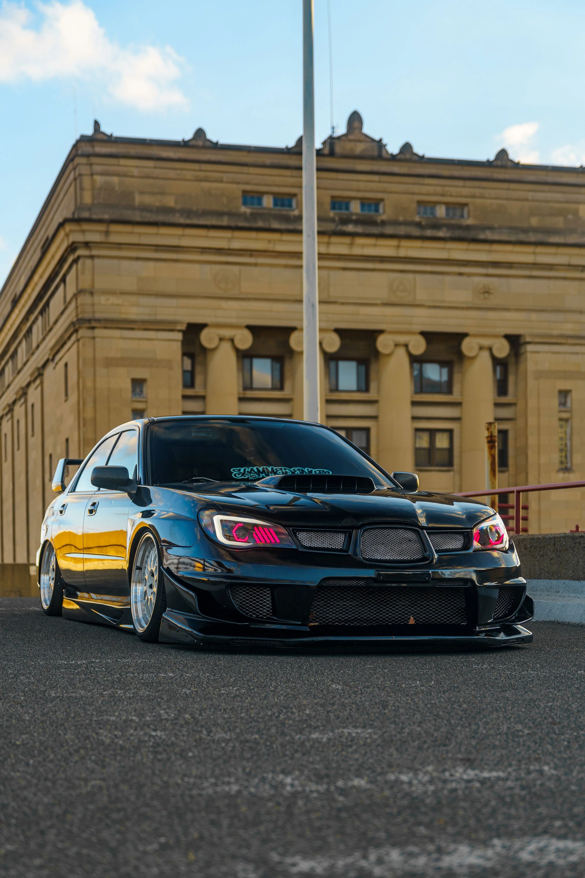 A black sporty car with custom pink lighting in the headlights is parked on a street in front of a historic building with columns and decorative stonework.