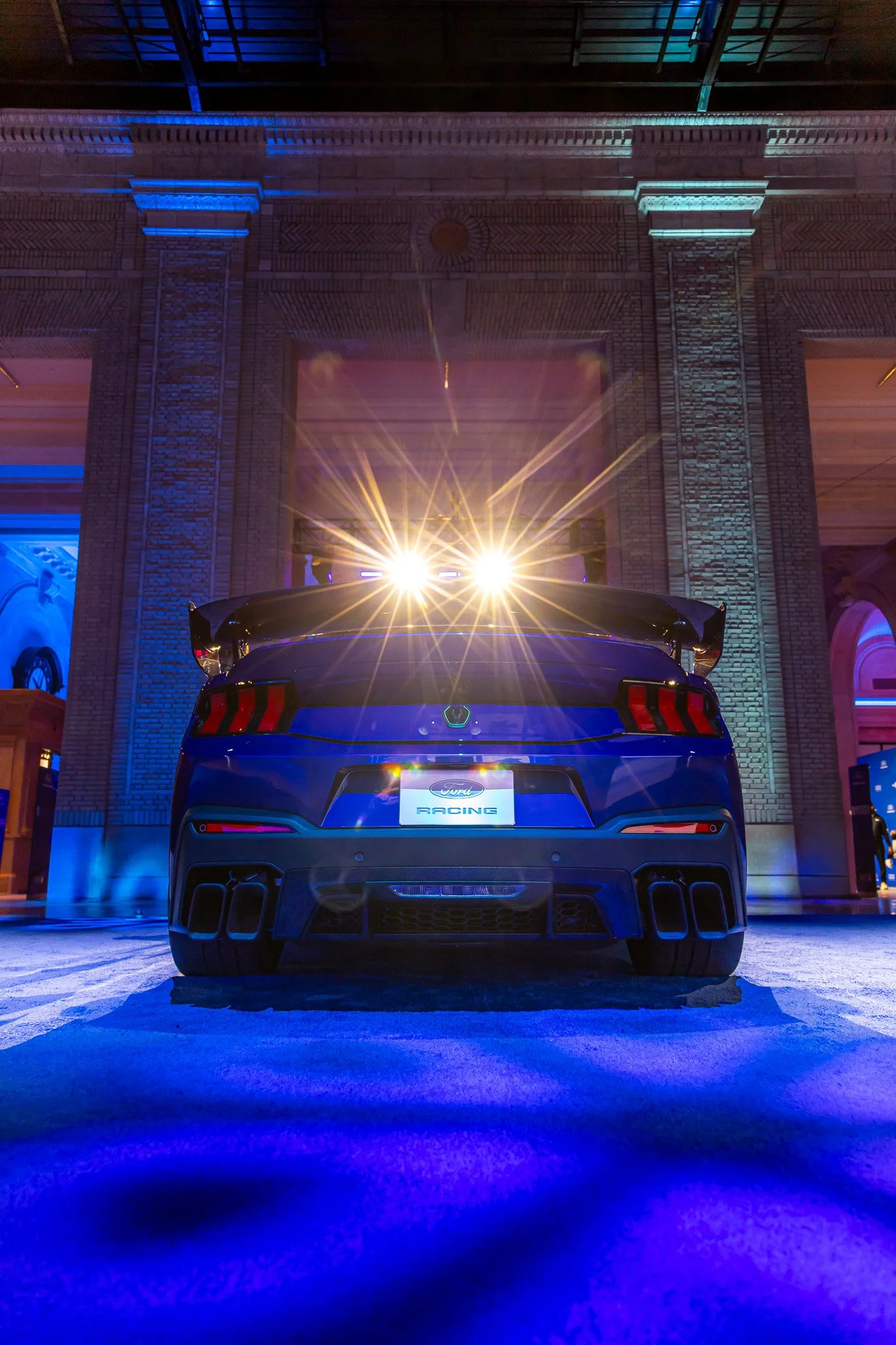 A blue Ford racing car with bright headlights parked inside a building with brick walls and blue and purple lighting.