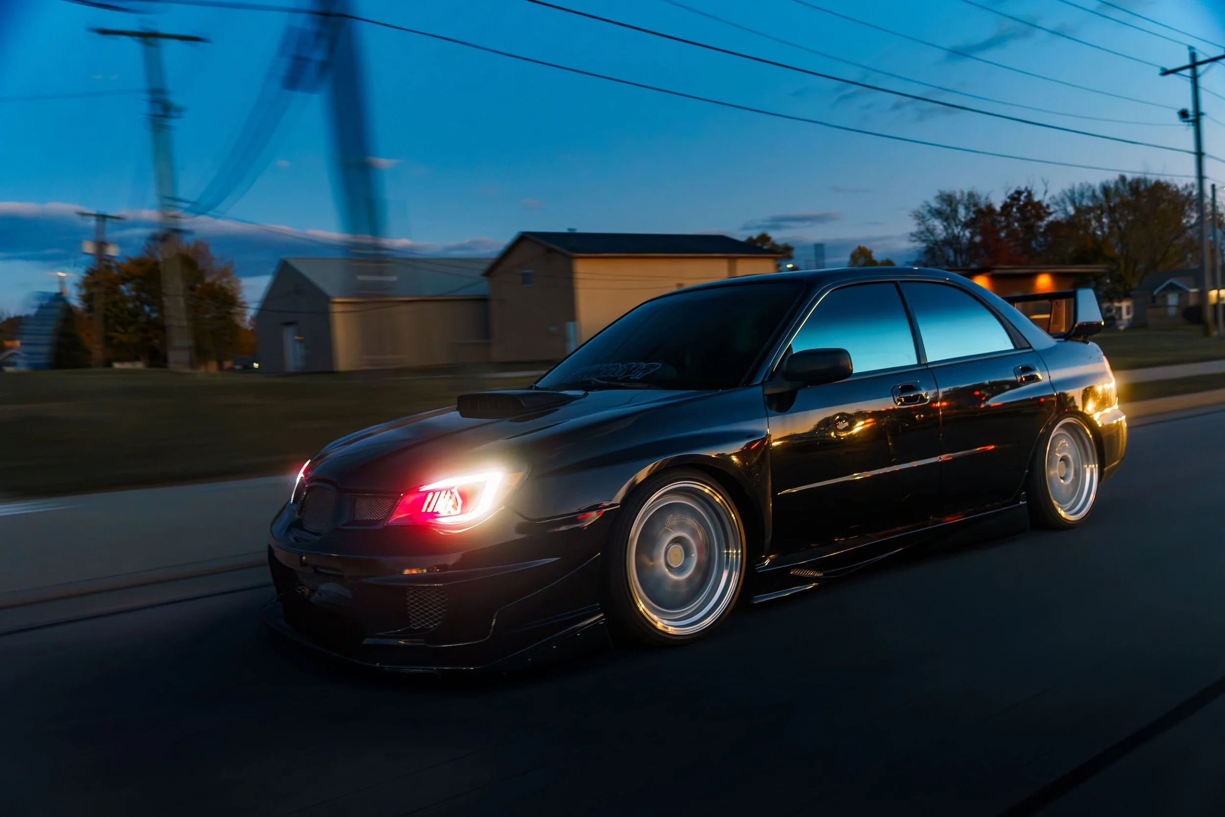 Black sports sedan driving on a road during dusk, with blurred background of houses and power lines.