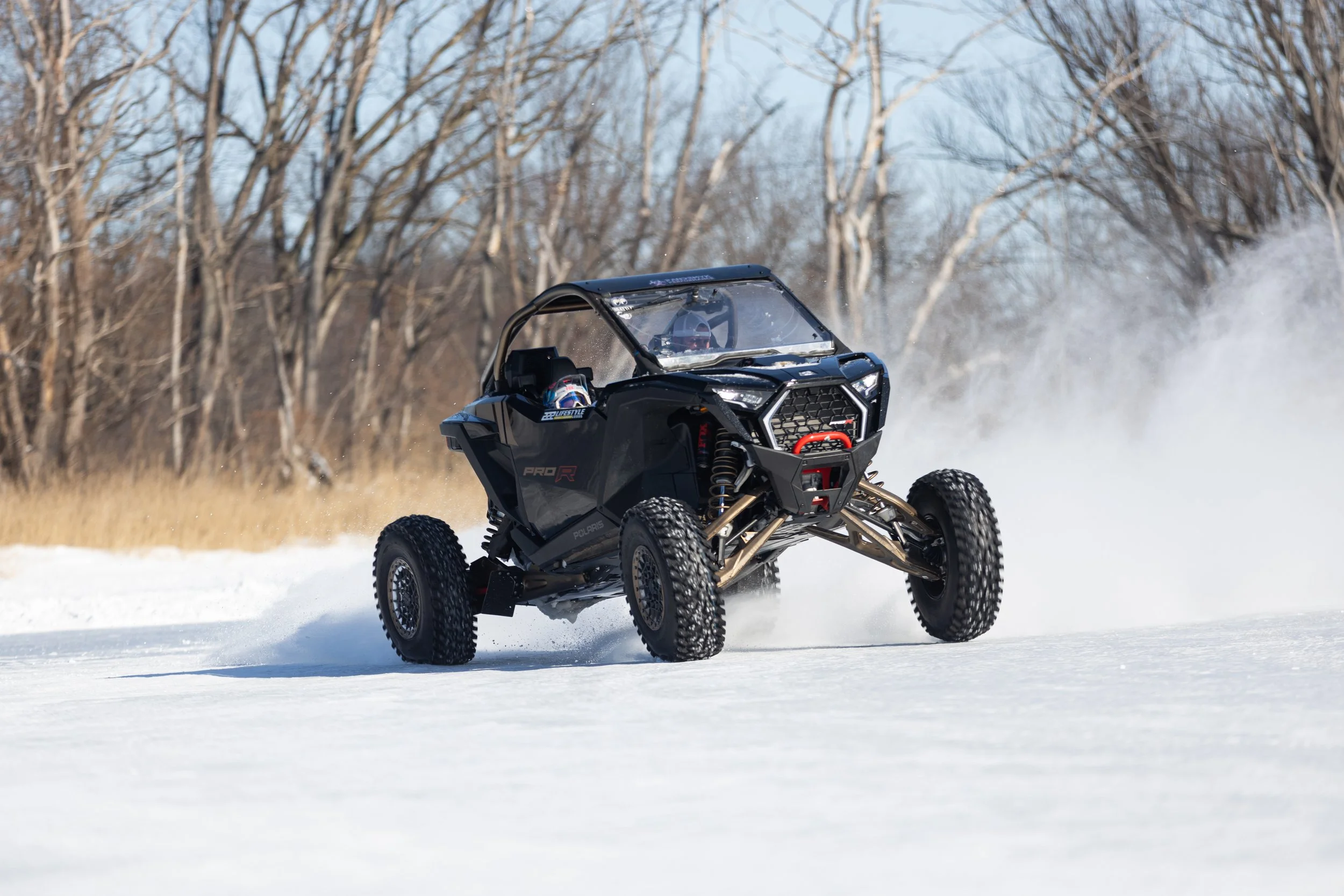 A Polaris off-road vehicle driving on snow-covered ground during winter with a background of leafless trees.