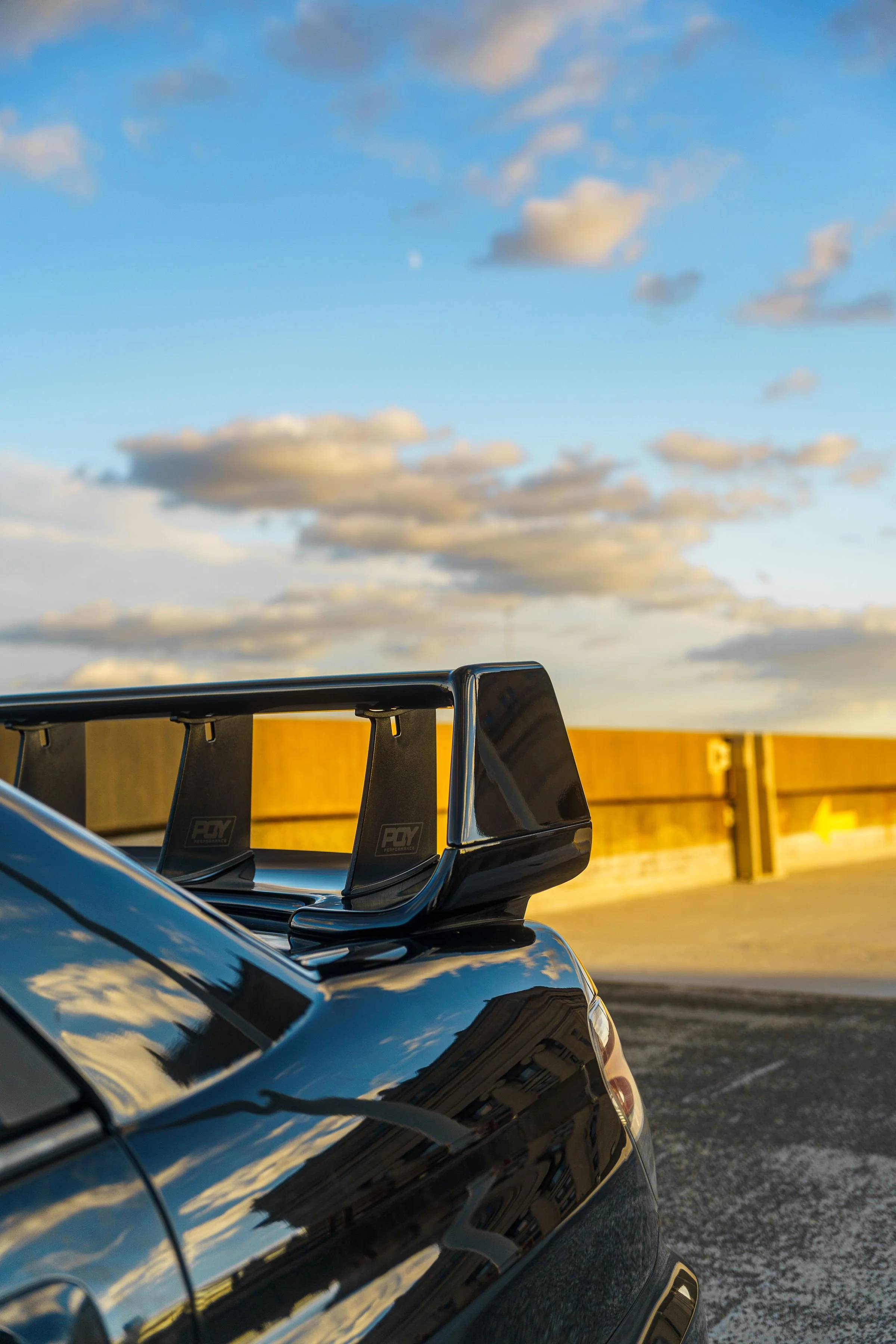 Close-up of a black car's rear with a large aerodynamic black rear wing spoiler, parked outdoors under a partly cloudy sky during sunset, reflecting the sky and the yellow barrier behind.