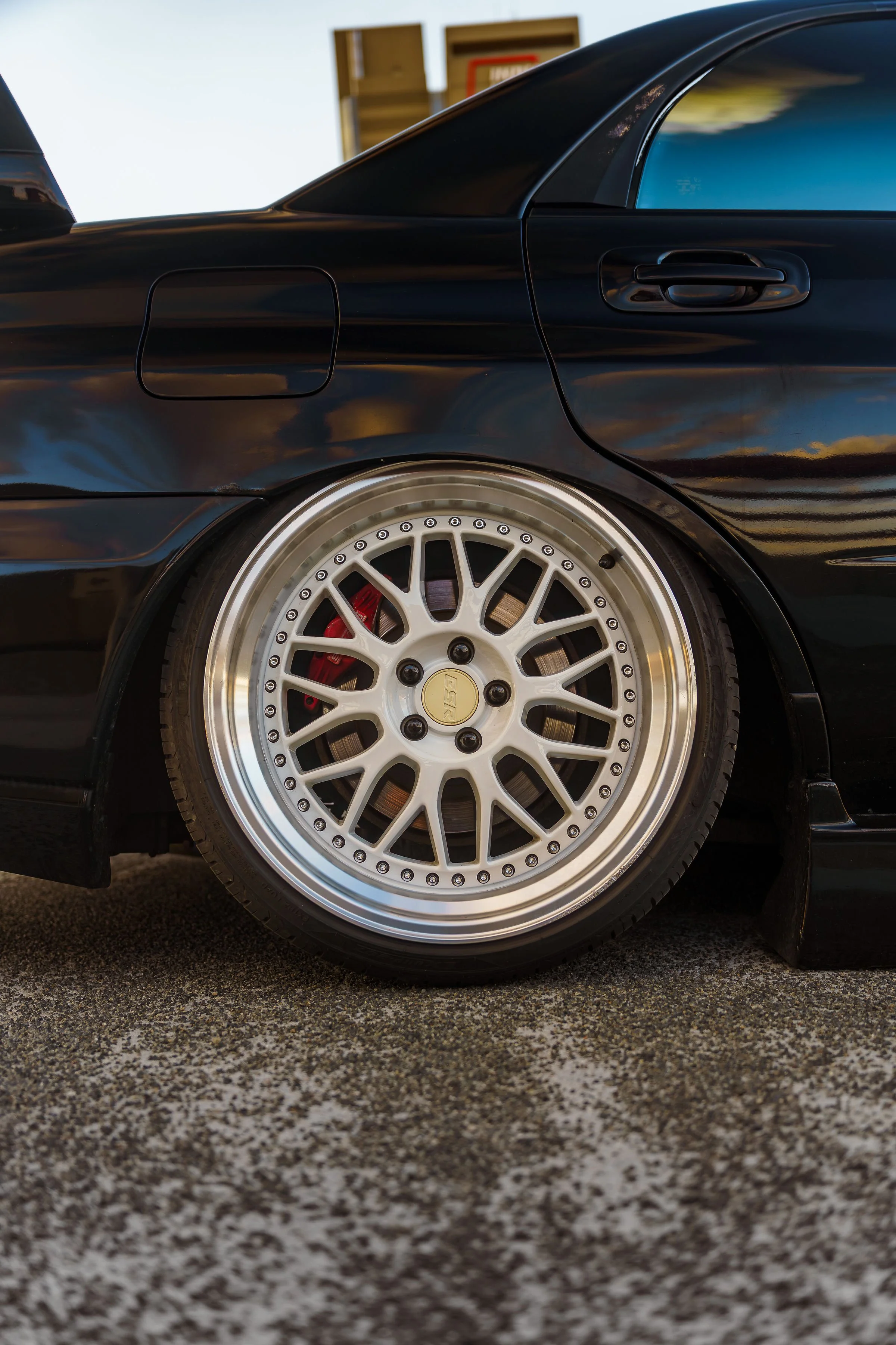 Close-up of a black sports car with a white multi-spoke wheel and red brake caliper, parked on textured pavement with a background of a building and blue sky.