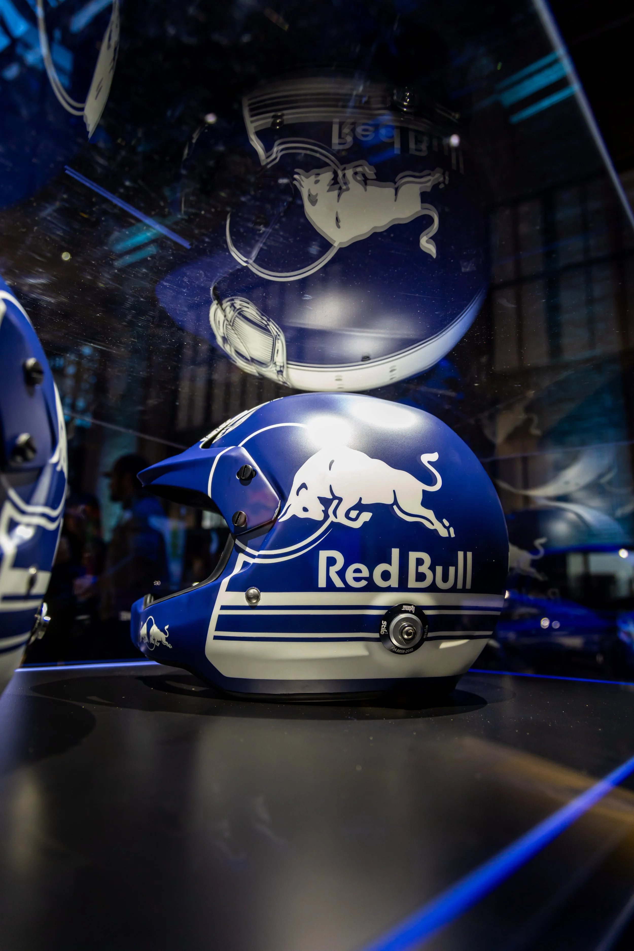 A blue Red Bull racing helmet with a white bull logo and the Red Bull logo displayed in a glass case.