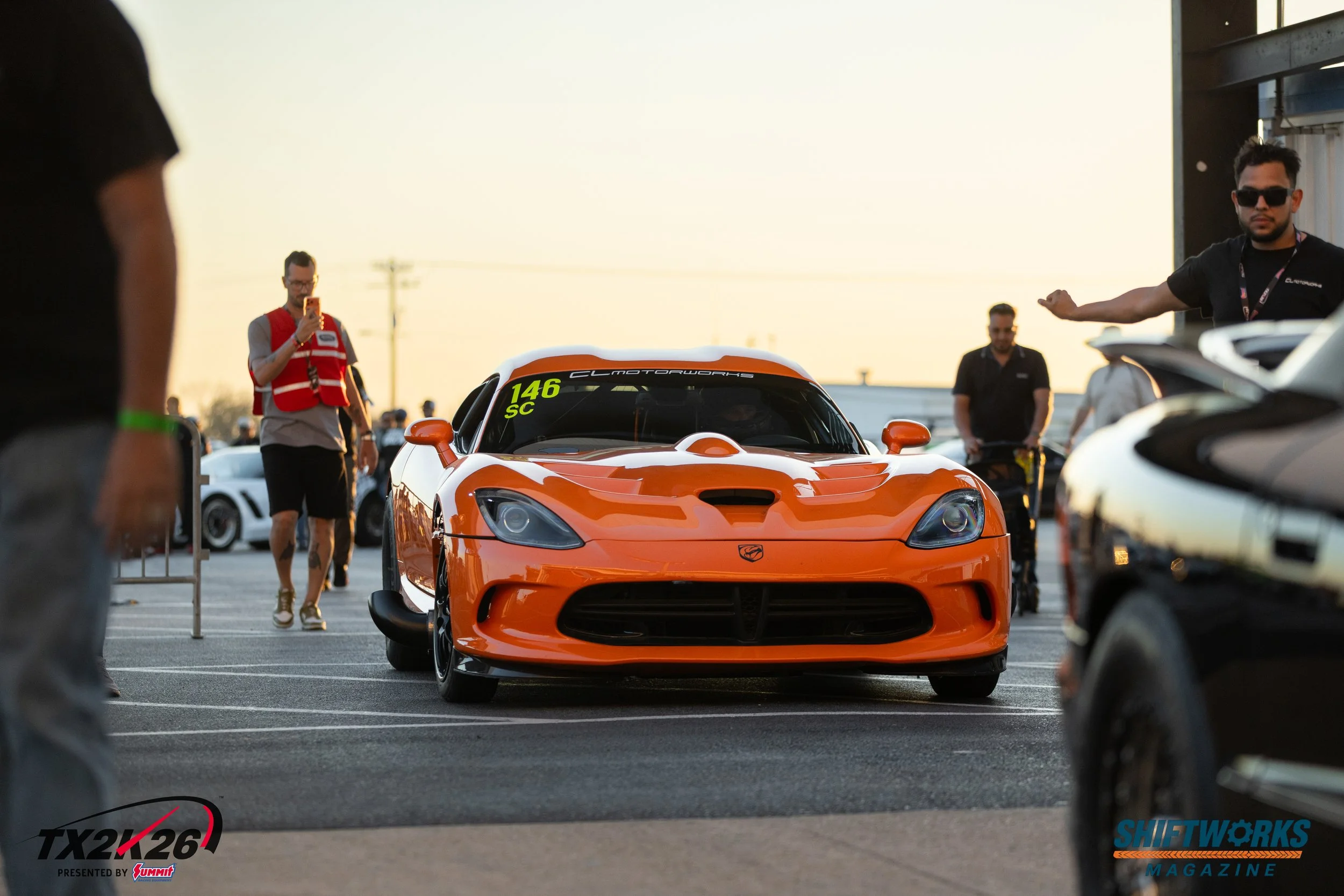 Orange sports car with black accents parked in a lot during sunset, surrounded by people, some taking photos and others gesturing, with logos of TX2K26, SHIFTW0RKS Magazine, and Summit visible on the image.