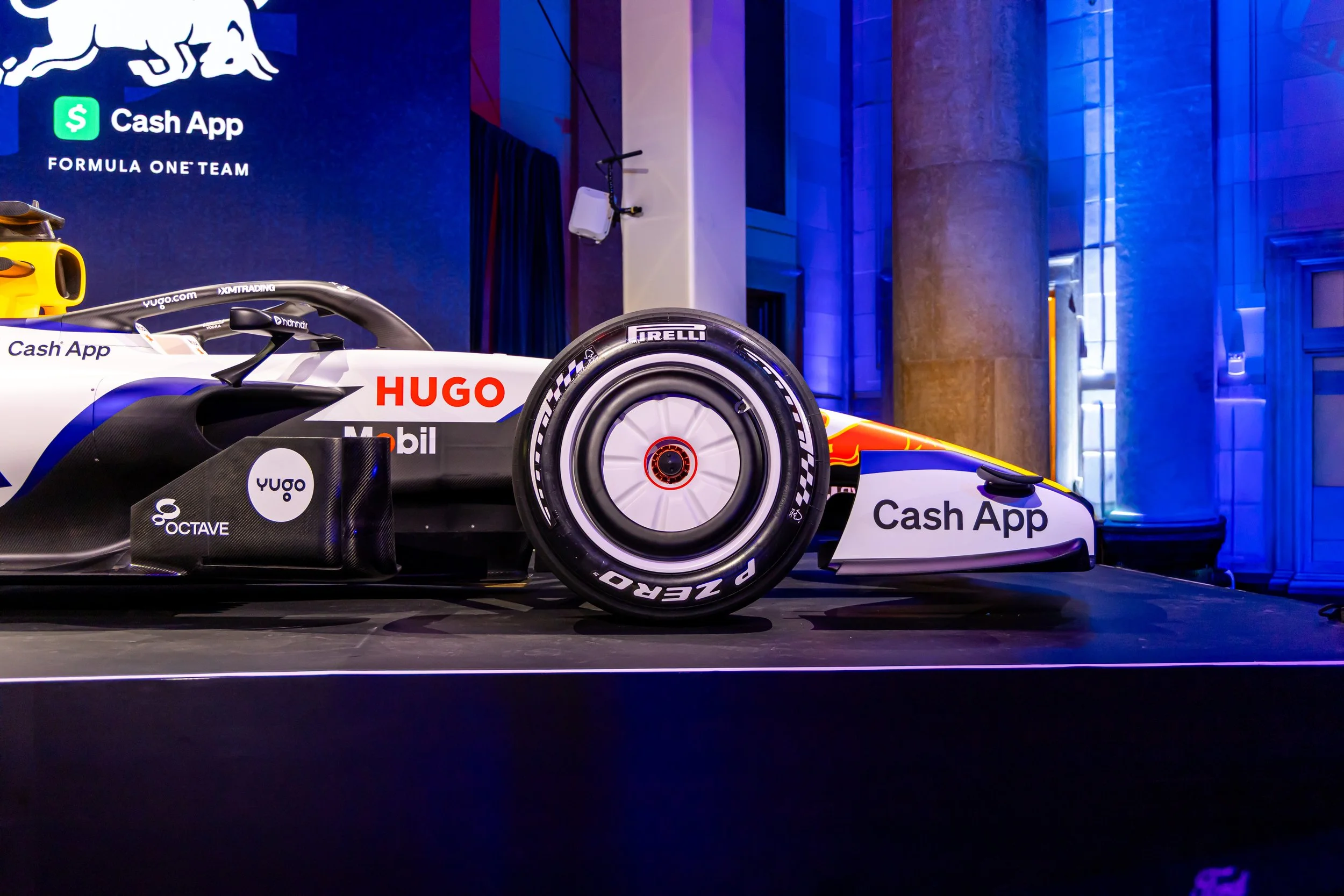 Formula 1 race car on display with sponsor logos, including Cash App, Hugo, and Pirelli, in a dimly lit room with blue lighting and pillars.