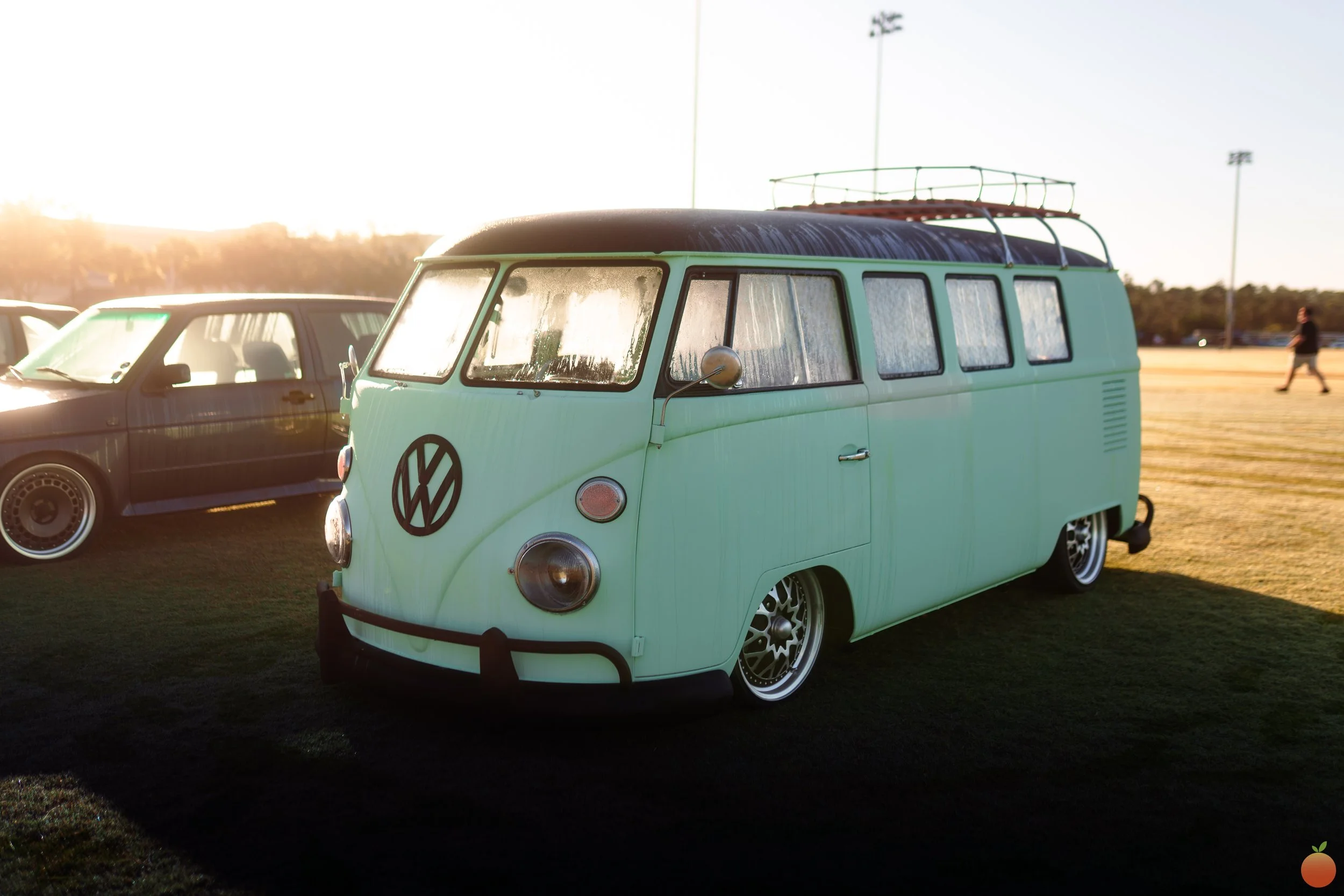 A vintage green Volkswagen camper van with a roof rack parked on a grassy field during sunset, with other cars and a person walking in the background.