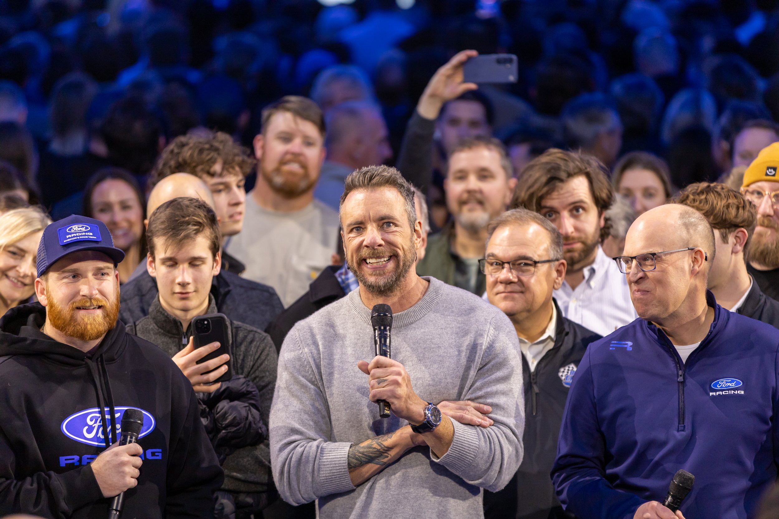 A man with a beard and gray sweater speaking into a microphone, surrounded by a diverse group of people at a public event or gathering.