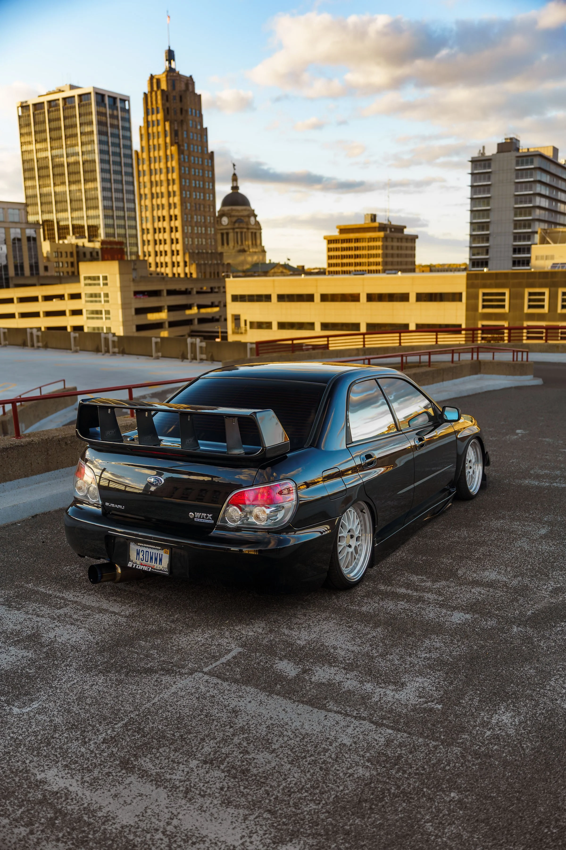 A black Subaru WRX car parked in a multi-level parking lot with a city skyline in the background, including tall buildings and a dome-shaped structure, during sunset.