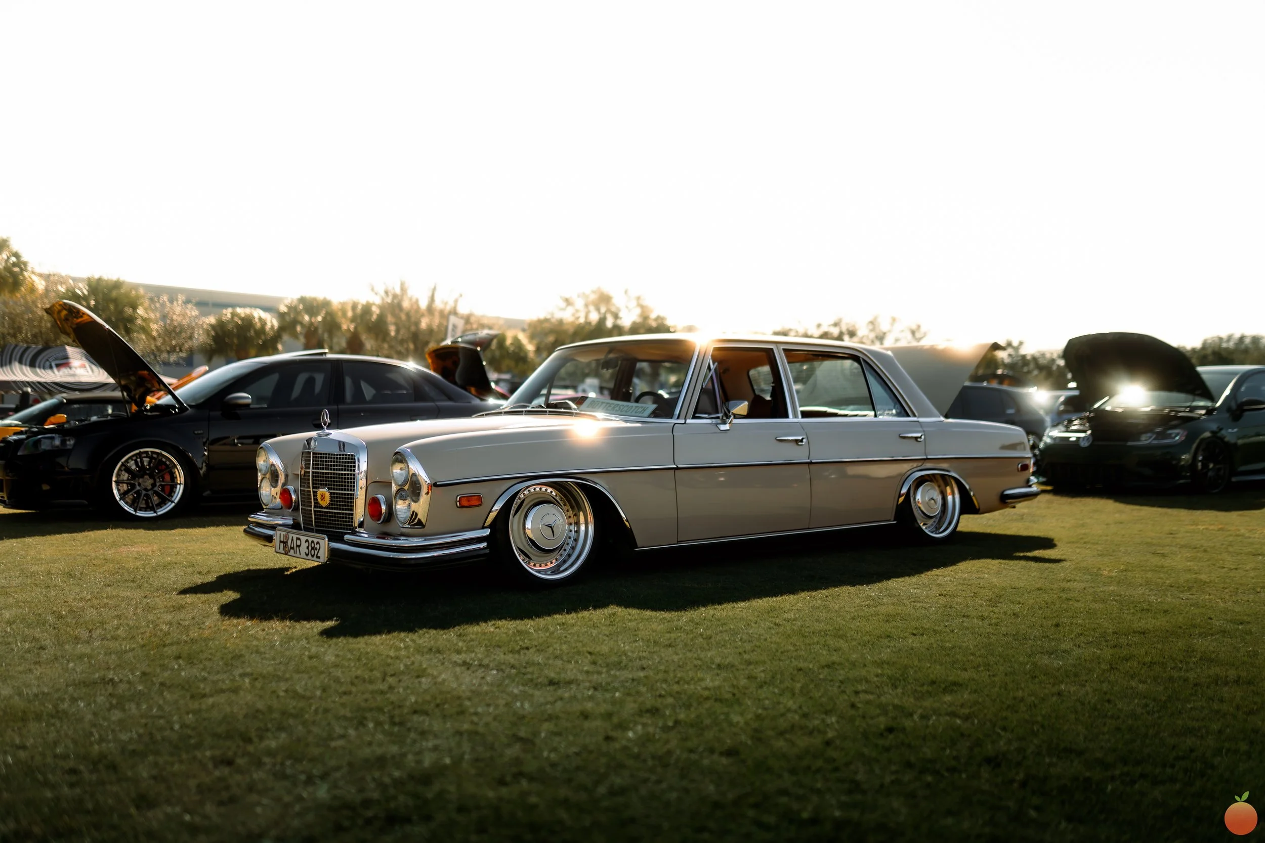 A vintage silver Mercedes-Benz car at a car show, parked on grass with other cars in the background, during sunset.