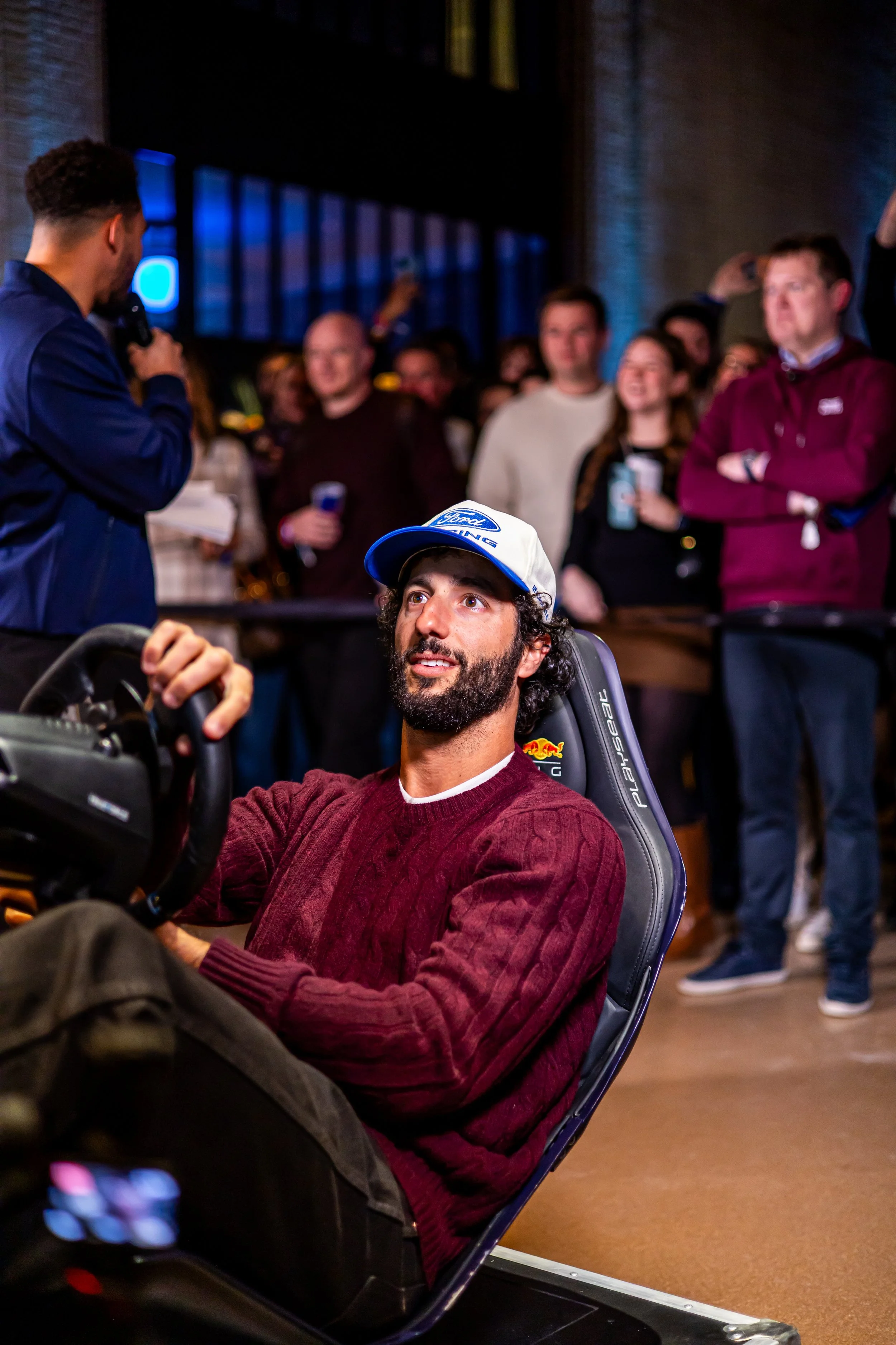 A man with a beard and curly hair wearing a burgundy sweater and a Ford racing cap sitting in a racing simulator chair, surrounded by a crowd of people at an indoor event.