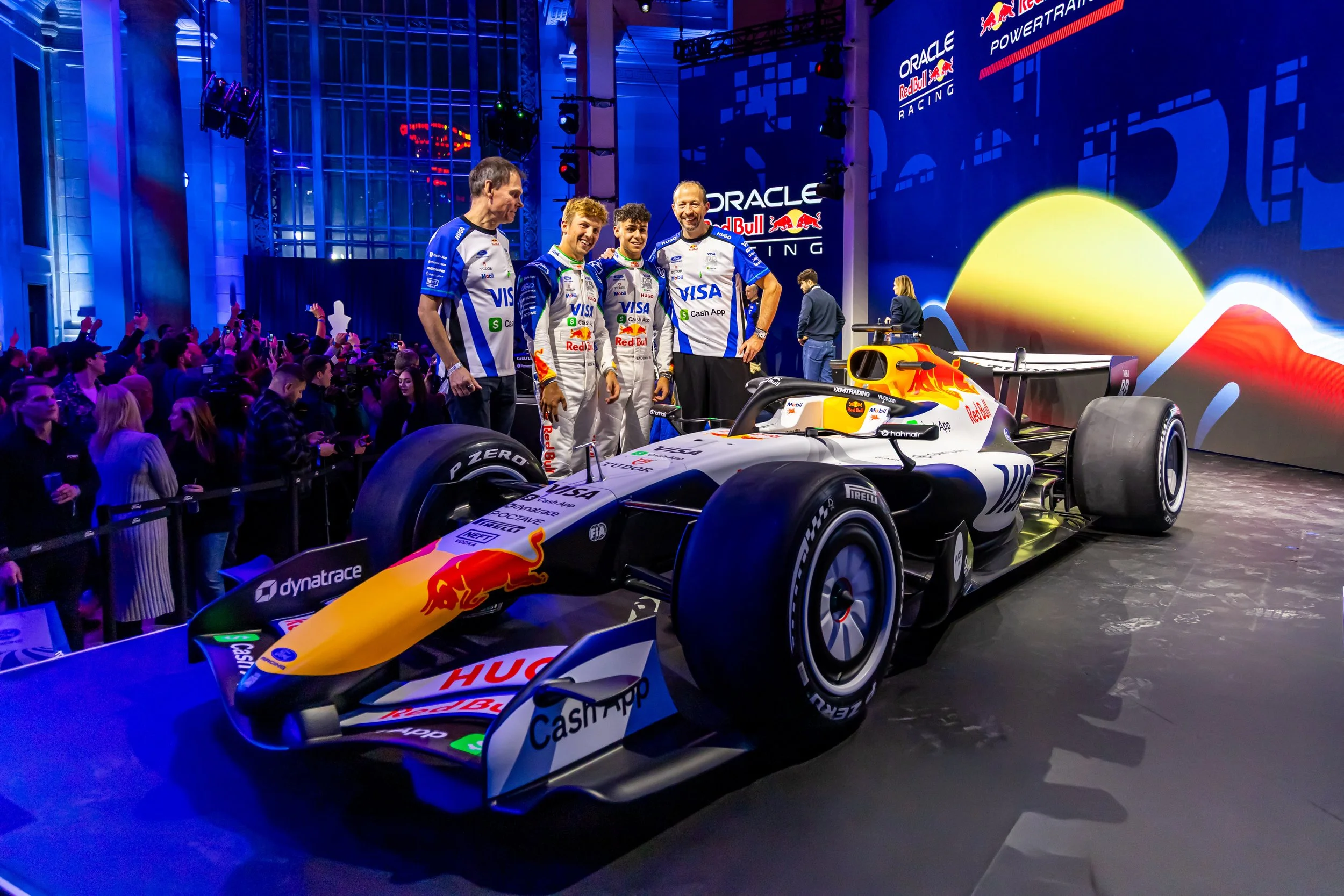 Four race car drivers in racing suits standing beside a Red Bull Racing Formula 1 car, with a large screen displaying the Red Bull logo in the background and an audience taking photos.