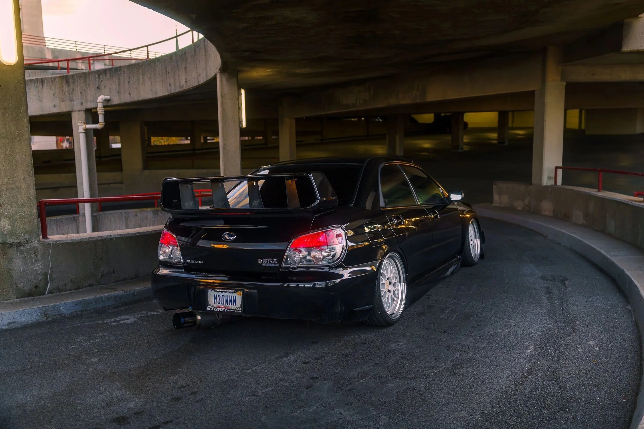 A black Subaru car with white rims parked in a parking garage underneath an overpass with concrete pillars and red guardrails.