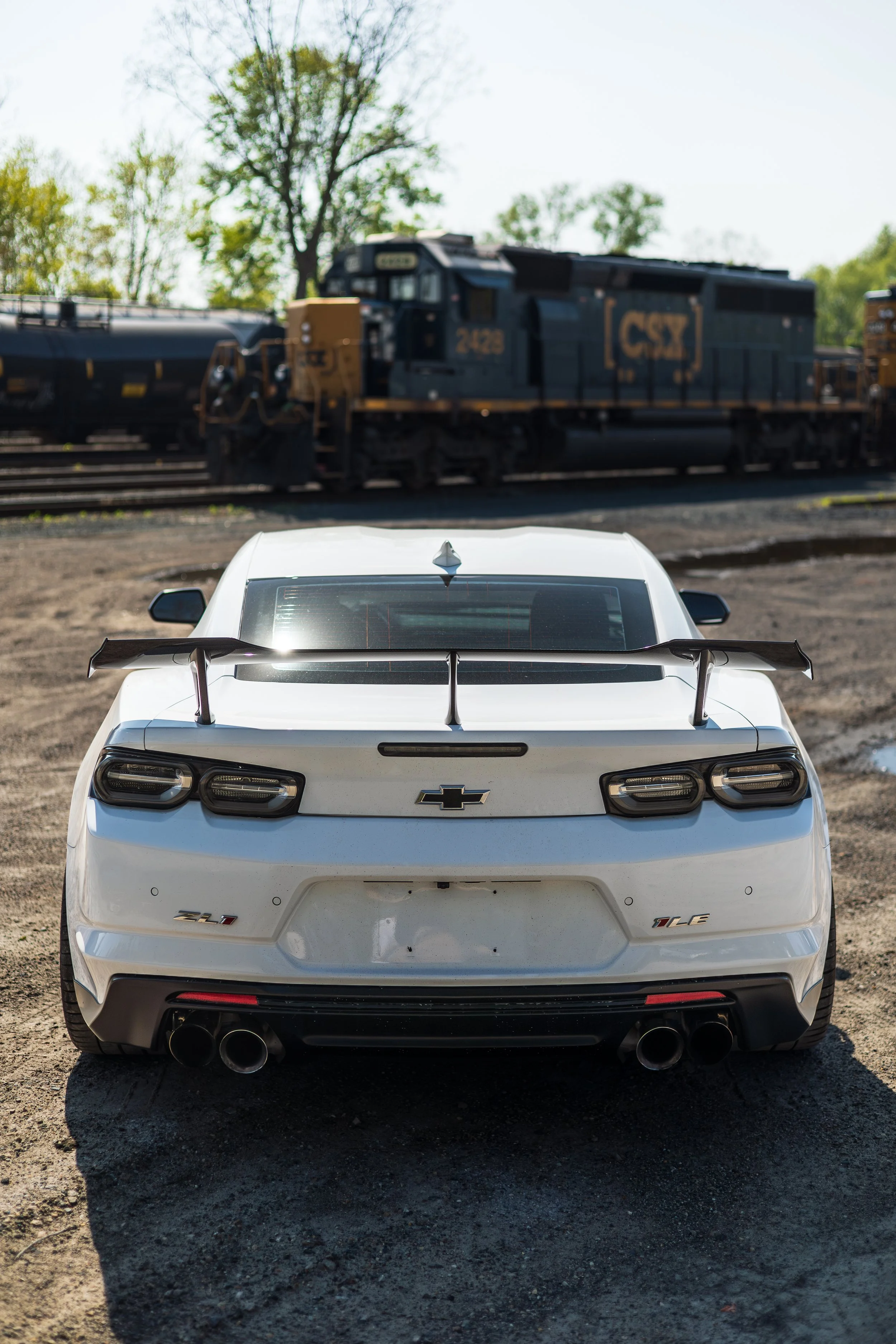 White Chevrolet Camaro ZL1 sports car with a large rear spoiler, parked on a dirt surface, with a black train locomotive in the background, trees, and clear sky.