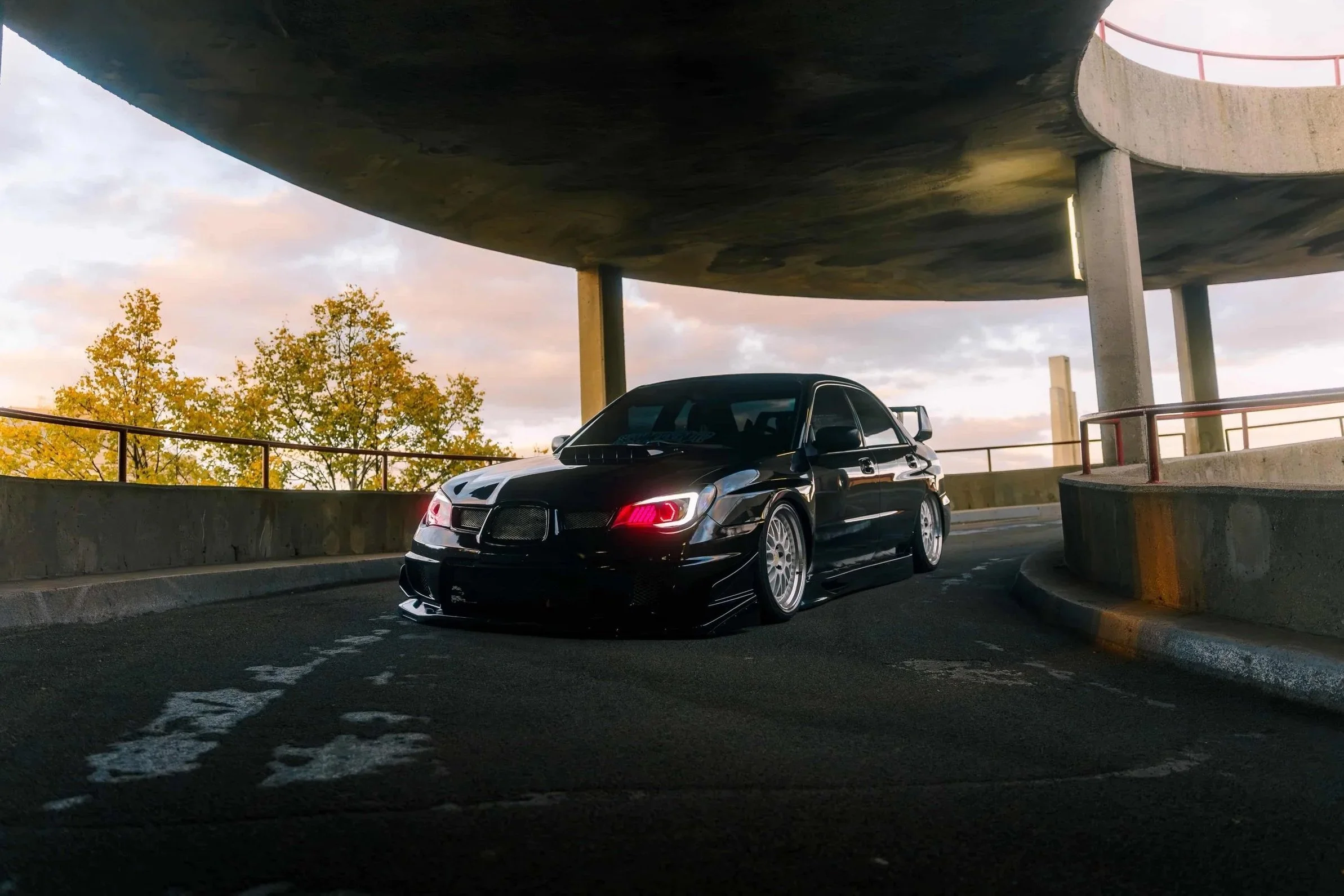 A black sports car parked in a circular parking garage under a concrete ceiling with trees and sky visible in the background.