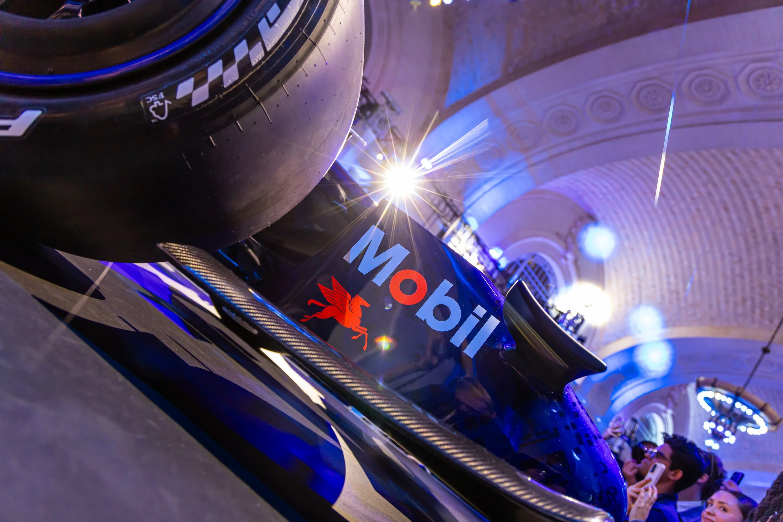 A racing car with Mobil branding on the side, situated in an indoor venue with ornate ceiling arches and blue lighting, spectators visible in the background.