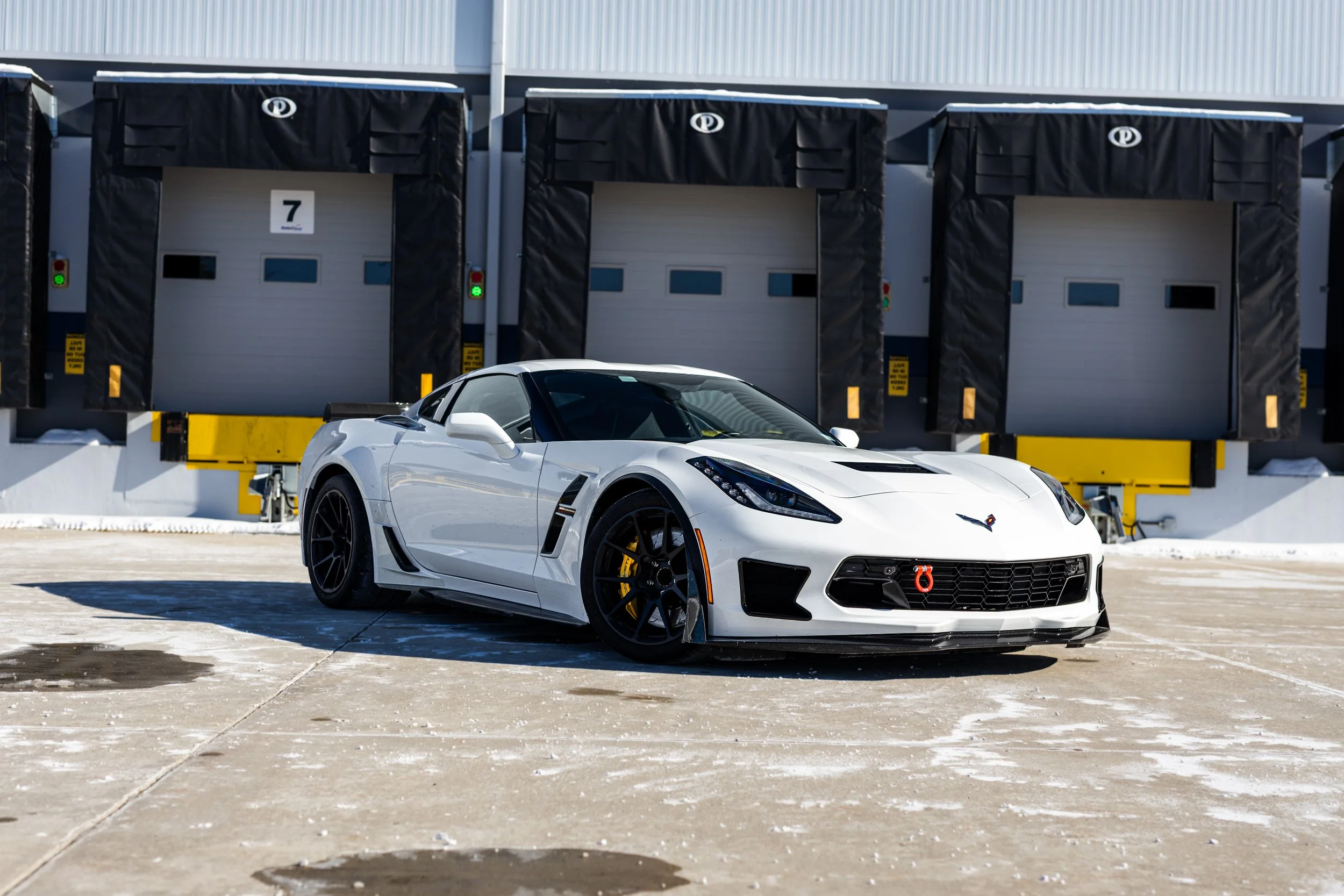 White sports car with black wheels parked in front of a industrial loading dock with three doors and black protective covers, on a sunny day with shadows and some snow on the ground.