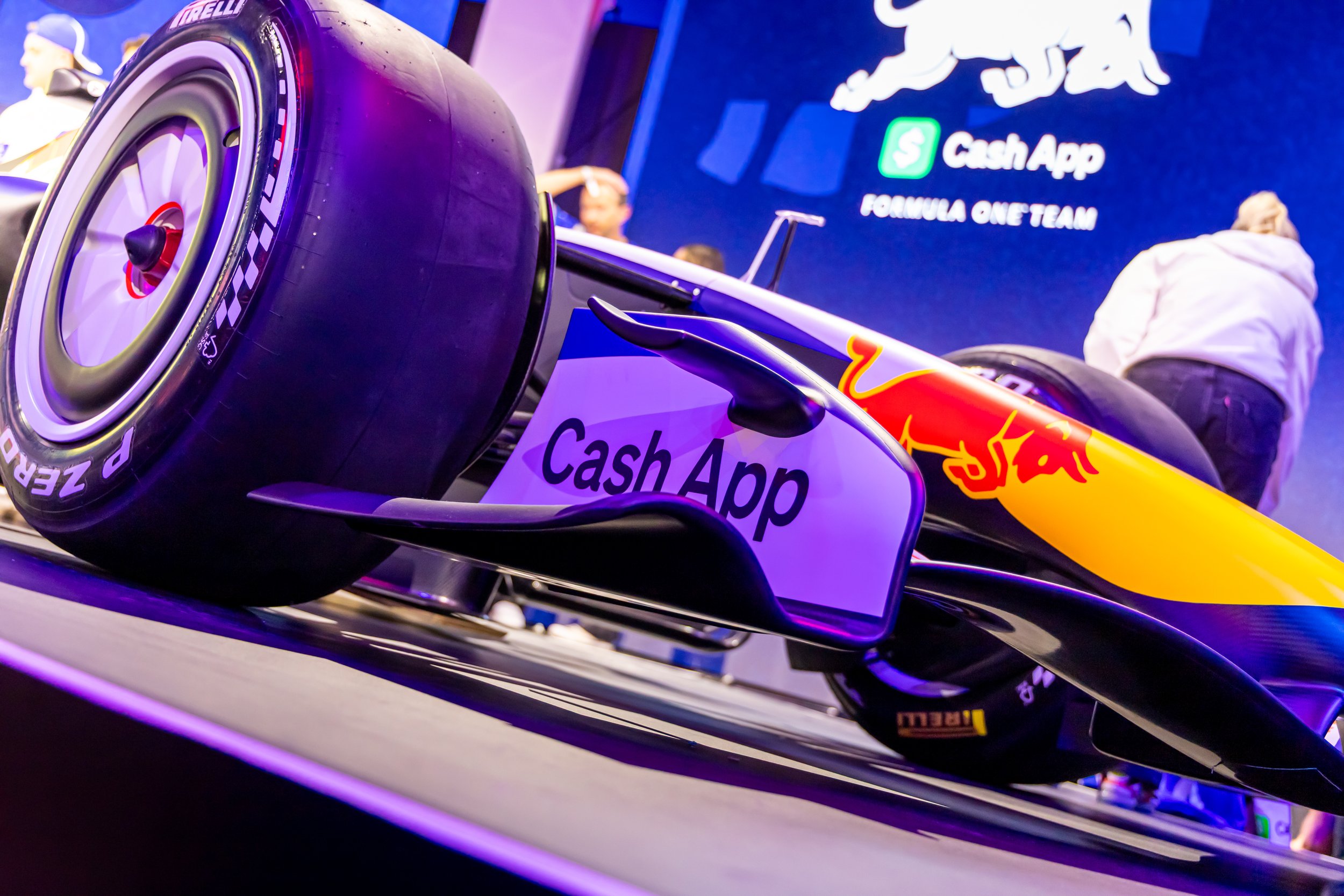 A Formula 1 race car with prominent Red Bull Racing branding and a Pirelli racing tire, displayed at an event with a large screen in the background showing the Cash App logo and the words 'Formula One Team.' People are seen in the background.