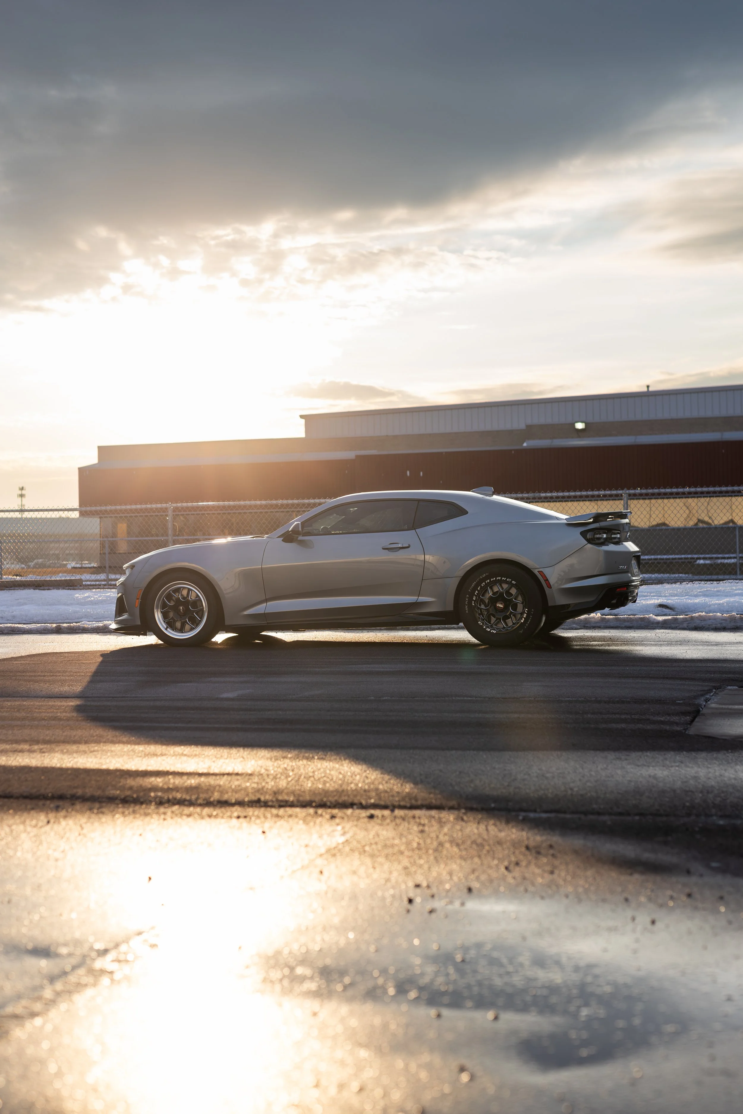 Silver car parked on a wet road during sunset with clouds in the sky.