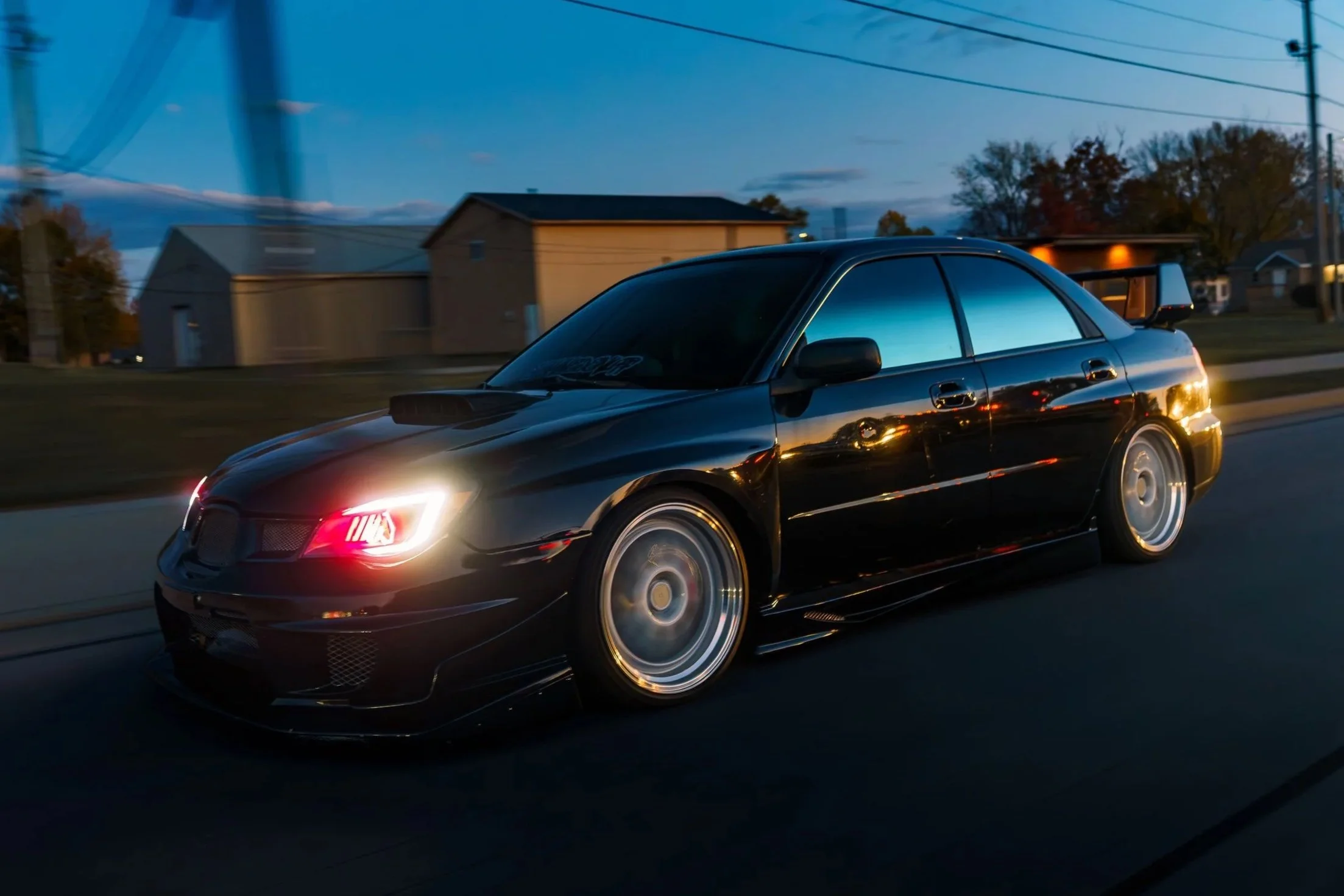 A black sports car with illuminated headlights driving on a road during dusk with houses and trees in the background.