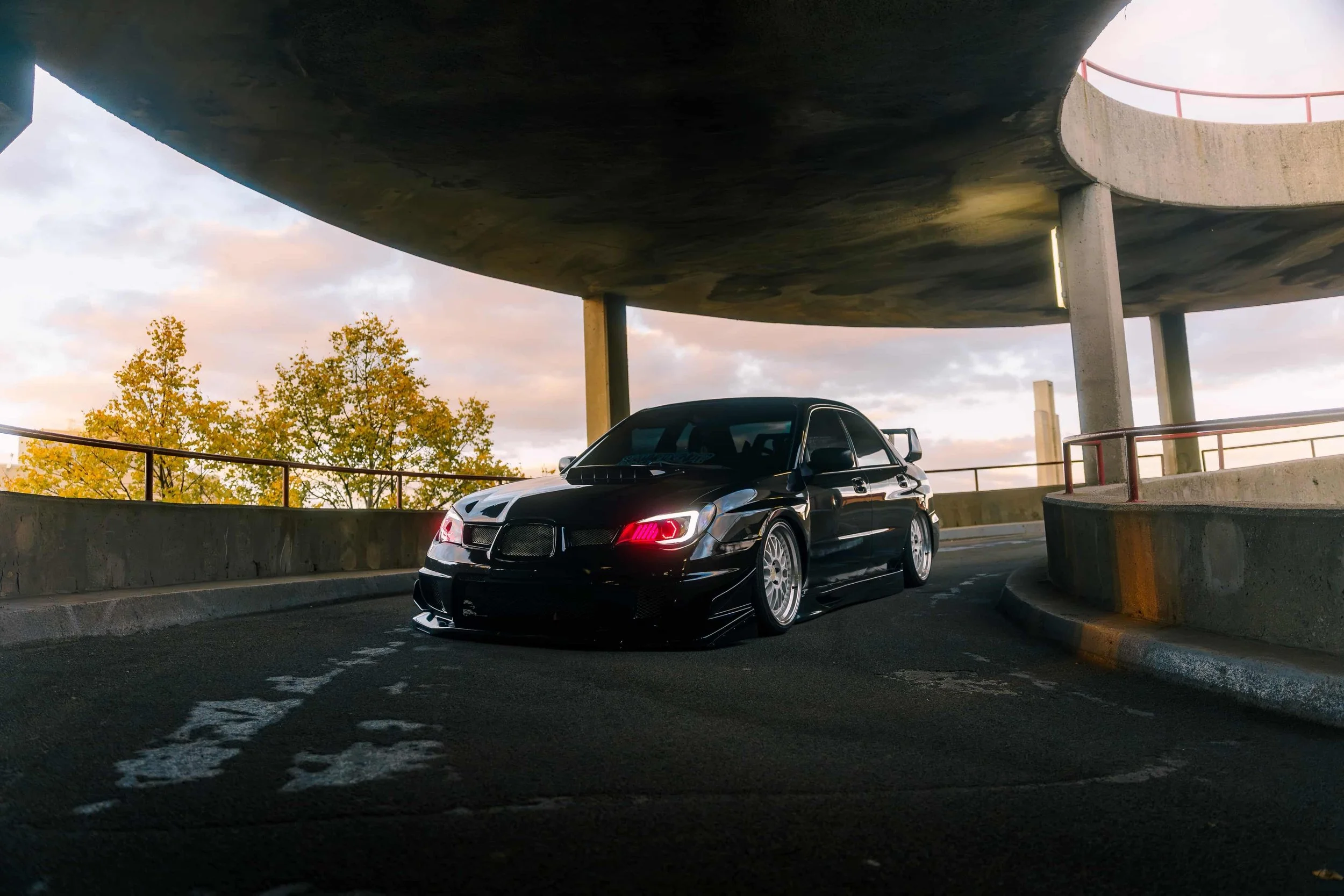 A black sports car parked under a circular concrete structure with trees and a cloudy sky in the background.