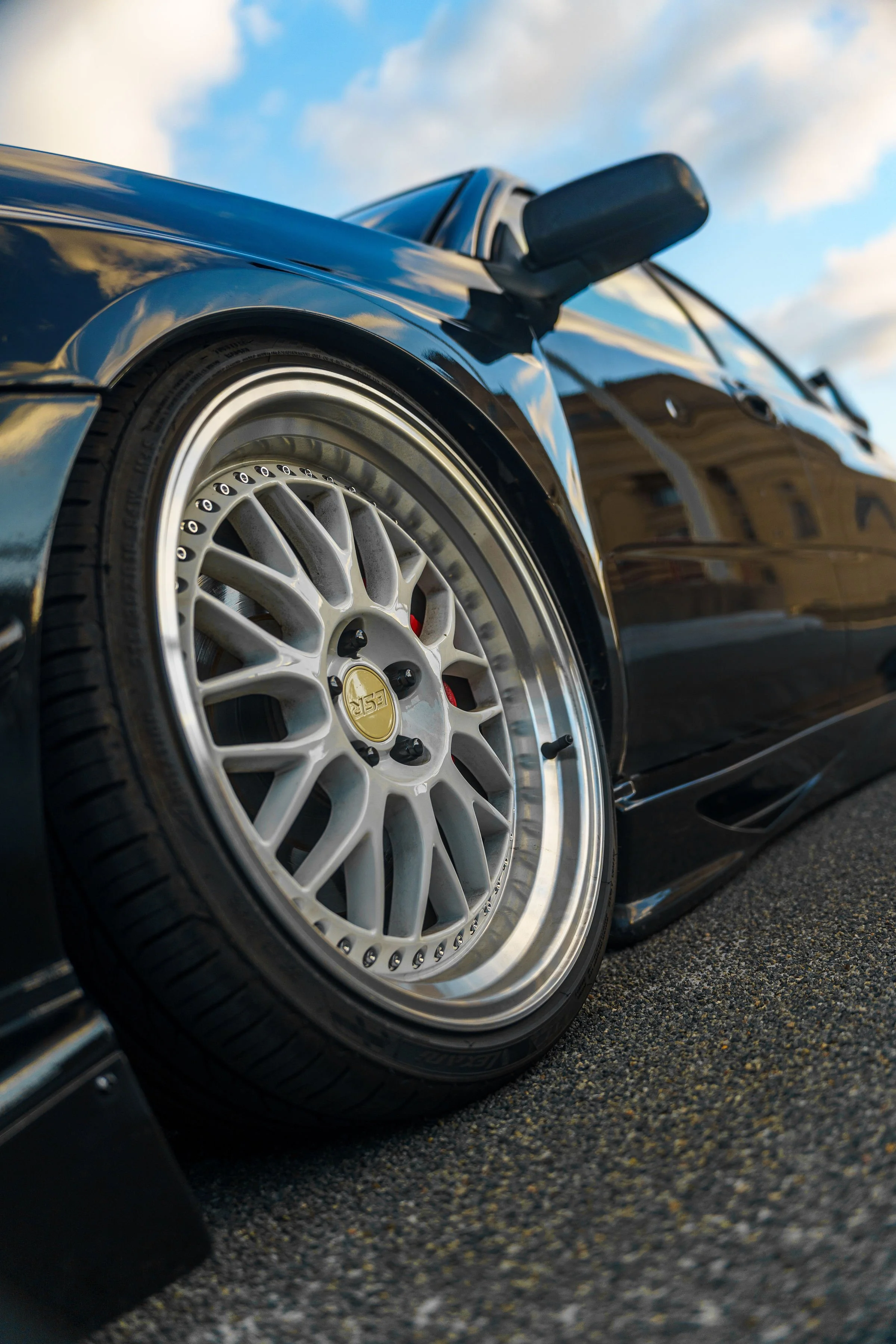Close-up of a black sports car's front left side with a white multi-spoke alloy wheel and a tire, reflecting the cloudy sky.