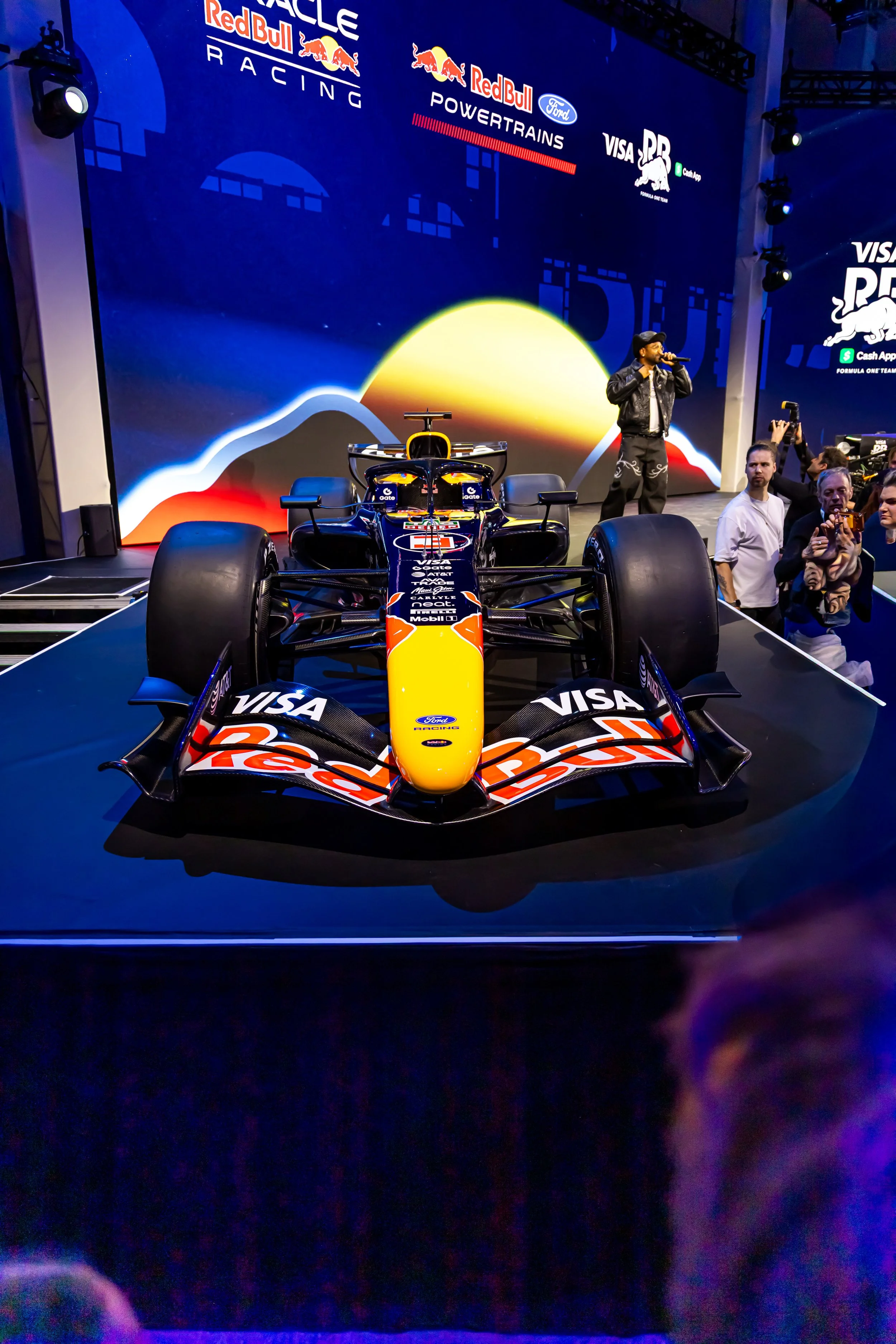 A Formula 1 race car with Red Bull Racing livery on display, with a large digital backdrop showing sunset graphics and the Red Bull Racing logo, at an indoor event.