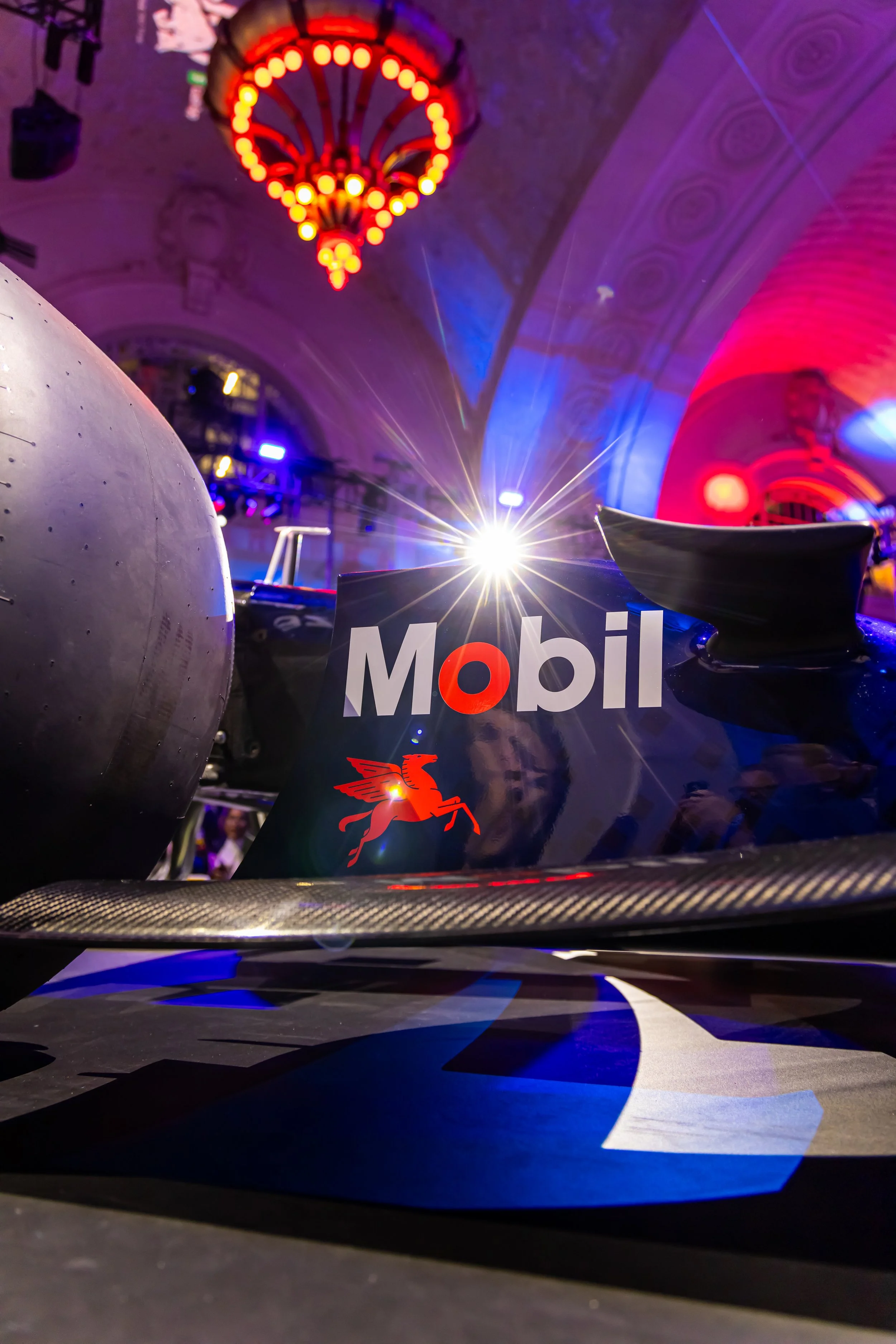 Close-up view of a Formula 1 race car with sponsor logos including Mobil and Chevalier, with a vibrant background of red and blue lighting and a chandelier hanging from the ceiling.
