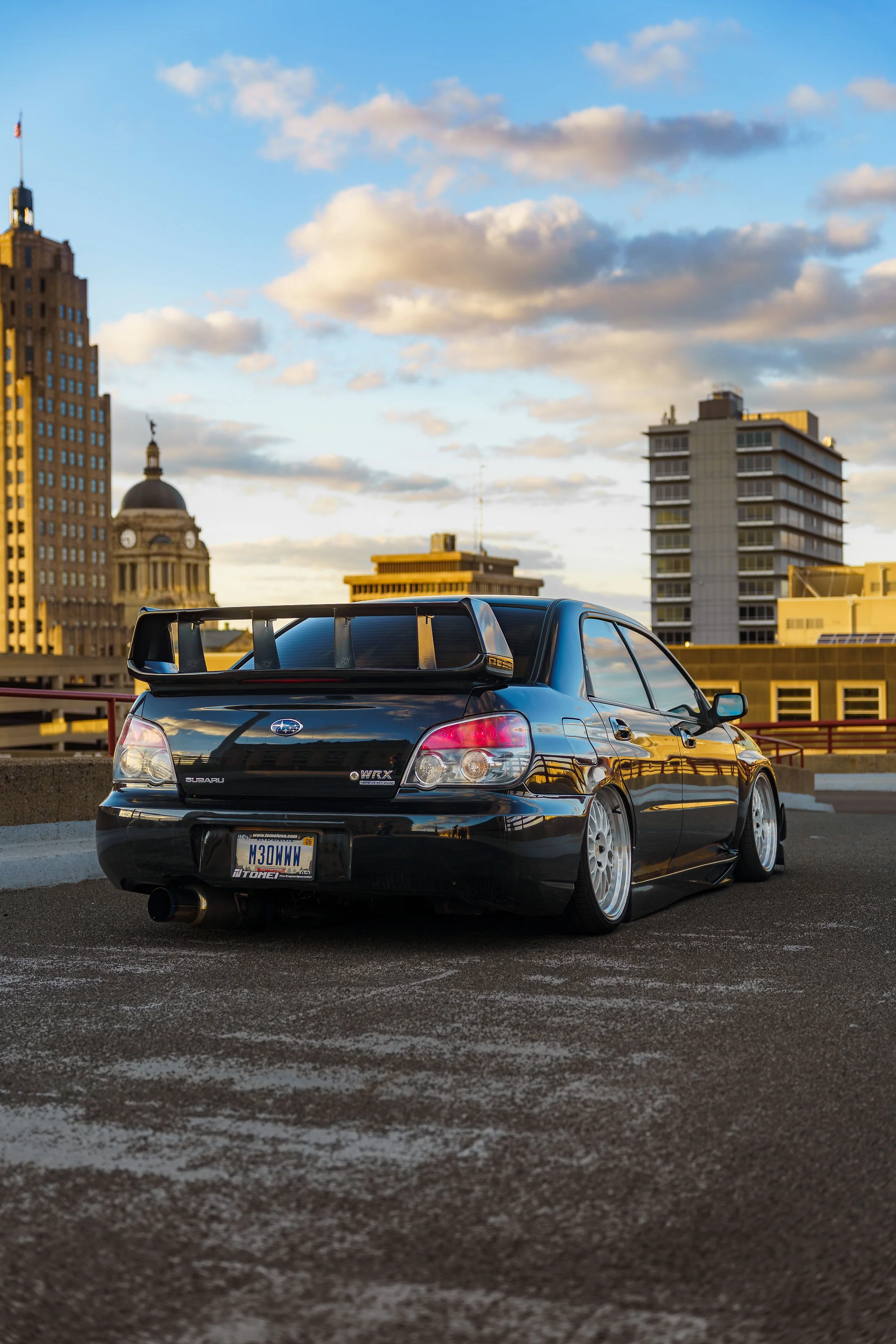A black Subaru WRX with a rear spoiler and aftermarket wheels is parked on a rooftop parking lot, with a city skyline featuring tall buildings and a partly cloudy sky in the background during the late afternoon or early evening.