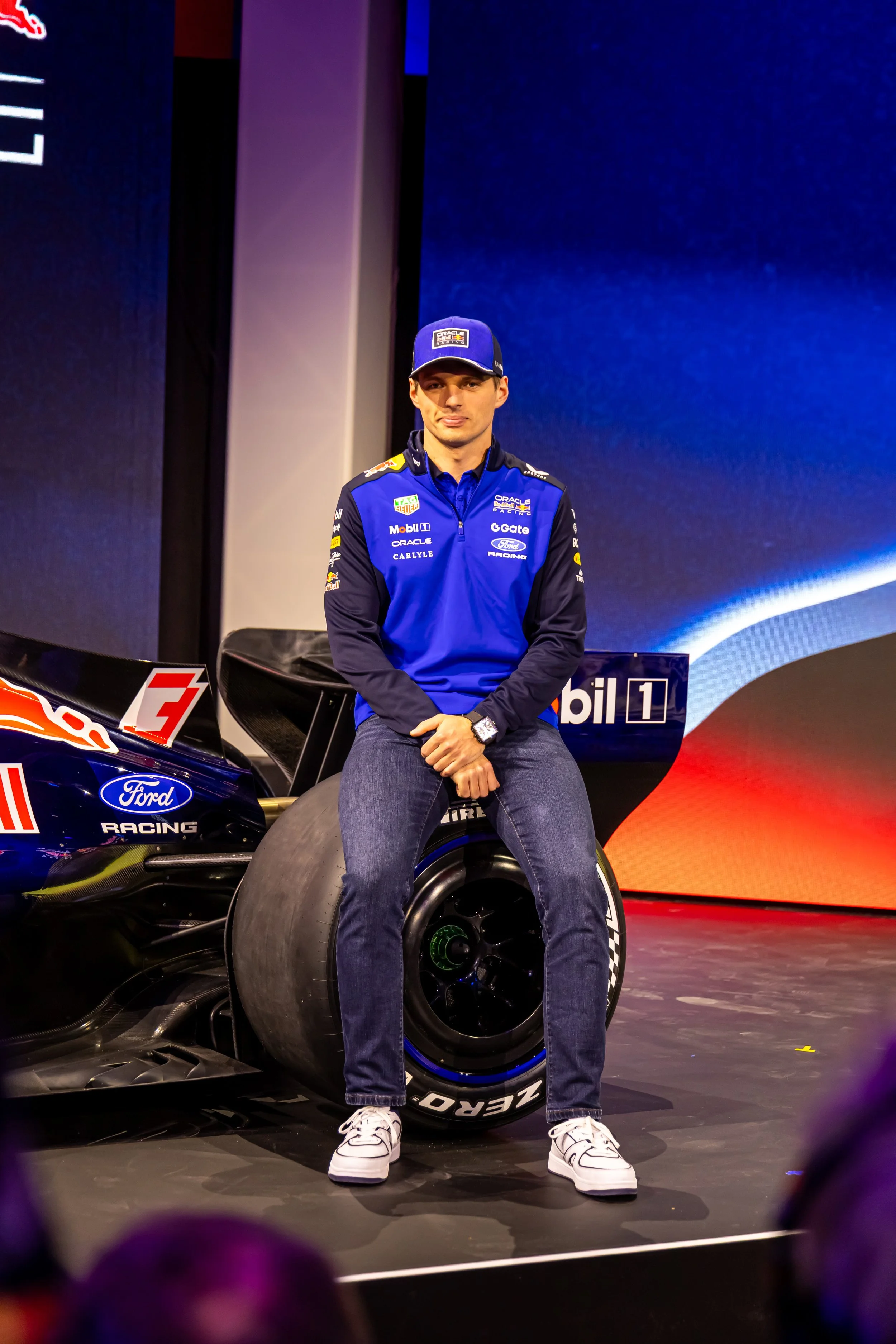 A man in a blue racing jacket and cap sits on a racing car's rear wing at an event.