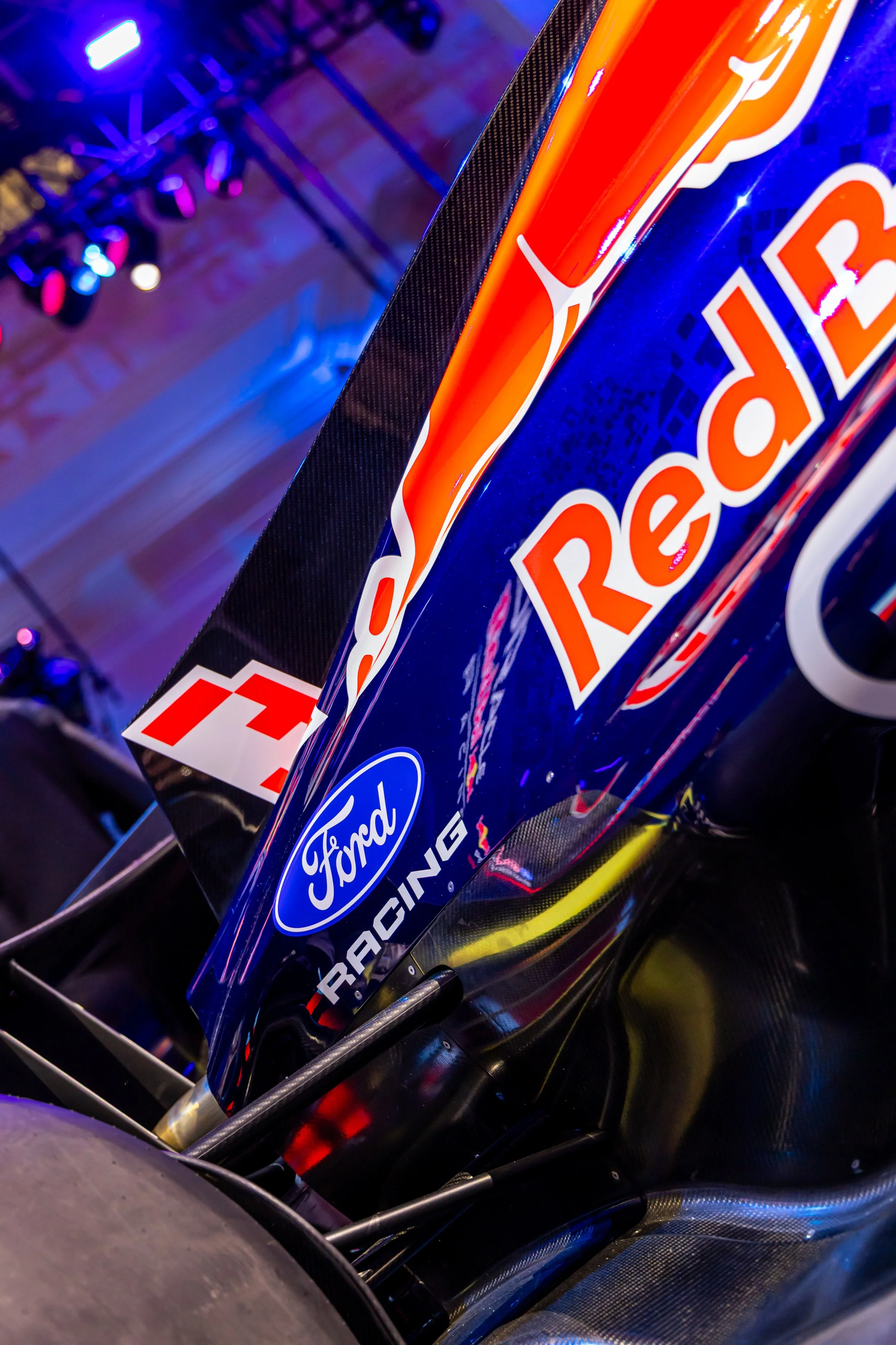Close-up of a Red Bull Racing Formula 1 car's rear wing with the Ford logo, captured in a dimly lit environment with colored stage lights.