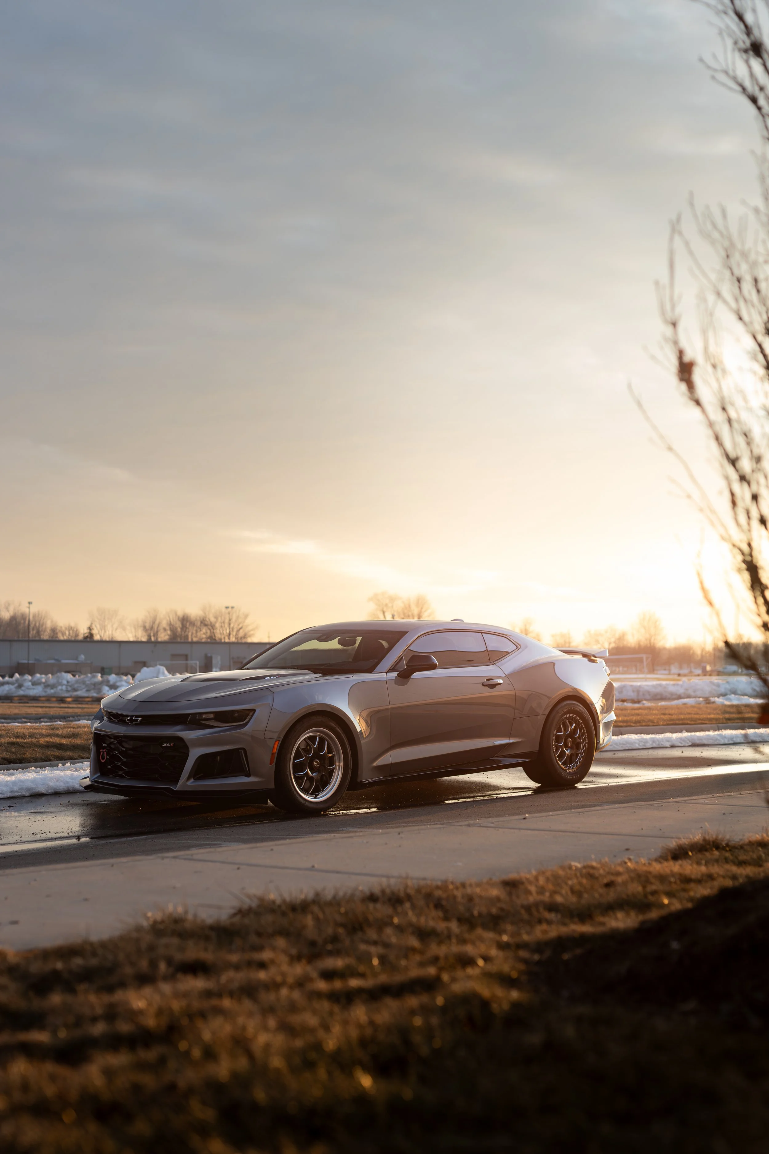A silver Chevrolet Camaro ZL1 parked on a wet driveway during sunset, with snow on the ground and a clear sky.