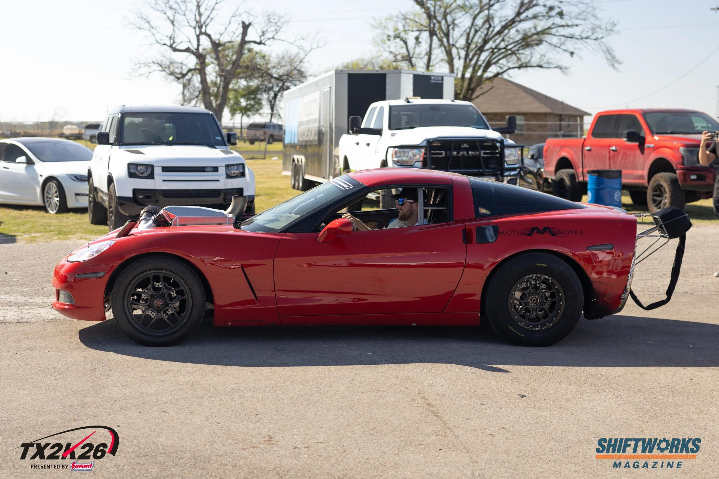 Red sports car with black wheels and a driver wearing sunglasses and a cap, parked at an outdoor event with other vehicles around, including trucks, an enclosed trailer, and a white car, under trees and a house in the background.