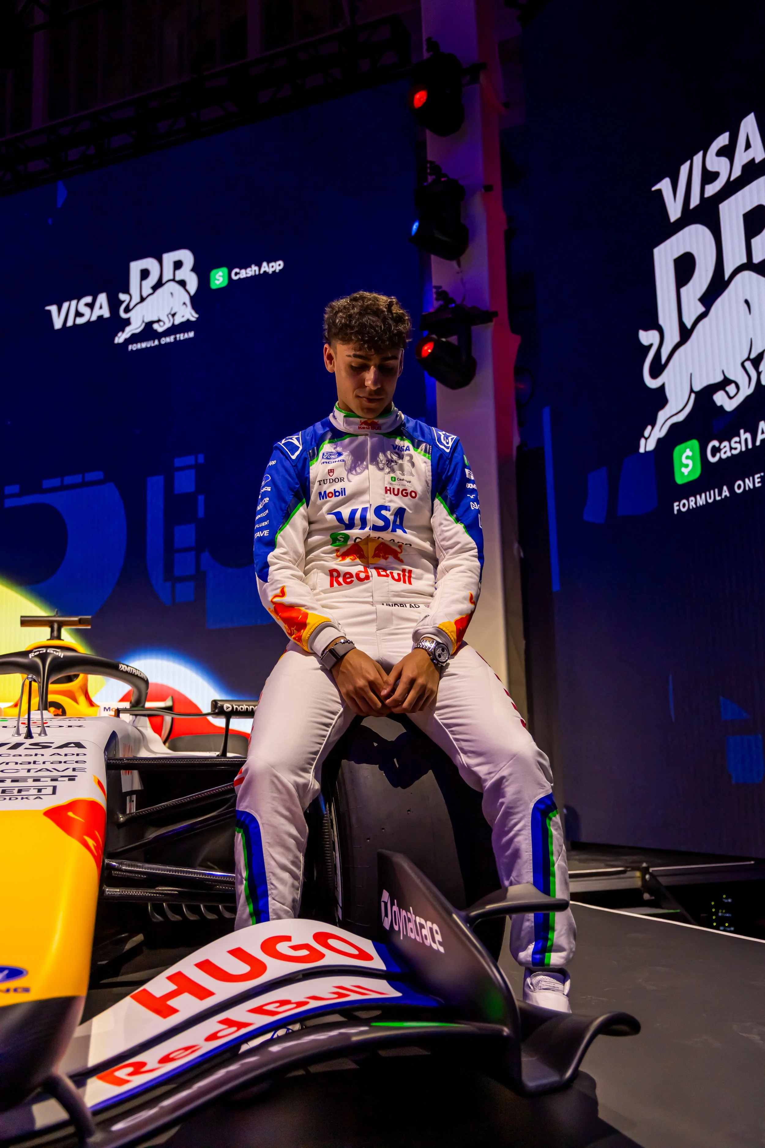 F1 driver sitting on a racing car in the garage during a race event, with sponsor logos on the car and a large digital screen in the background.