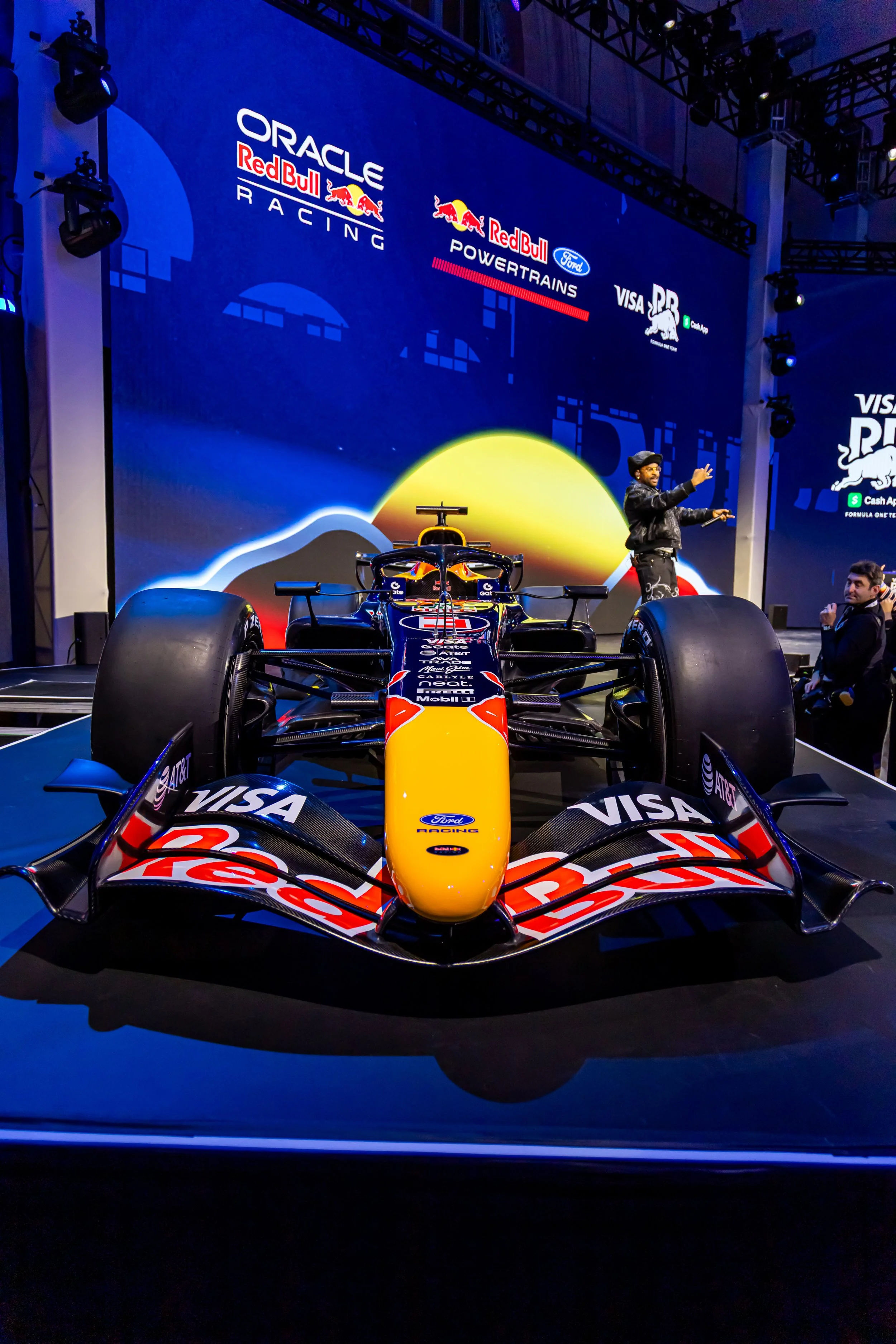 Formula 1 race car displayed on stage with Red Bull branding, large screen in background showing logos of Oracle Red Bull Racing, Red Bull Ford Powertrains, and Visa, with a sunset or sunrise graphic.