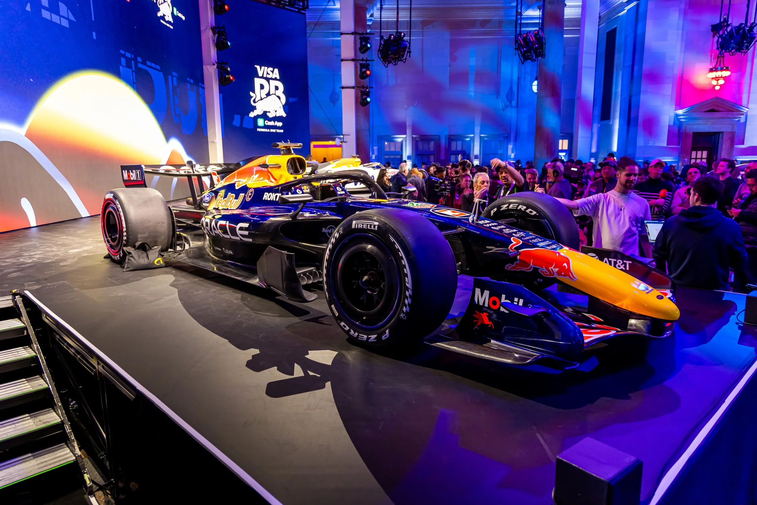 A Formula 1 race car displayed on a stage at an indoor event, surrounded by a crowd of people, with colorful lighting and graphics in the background.