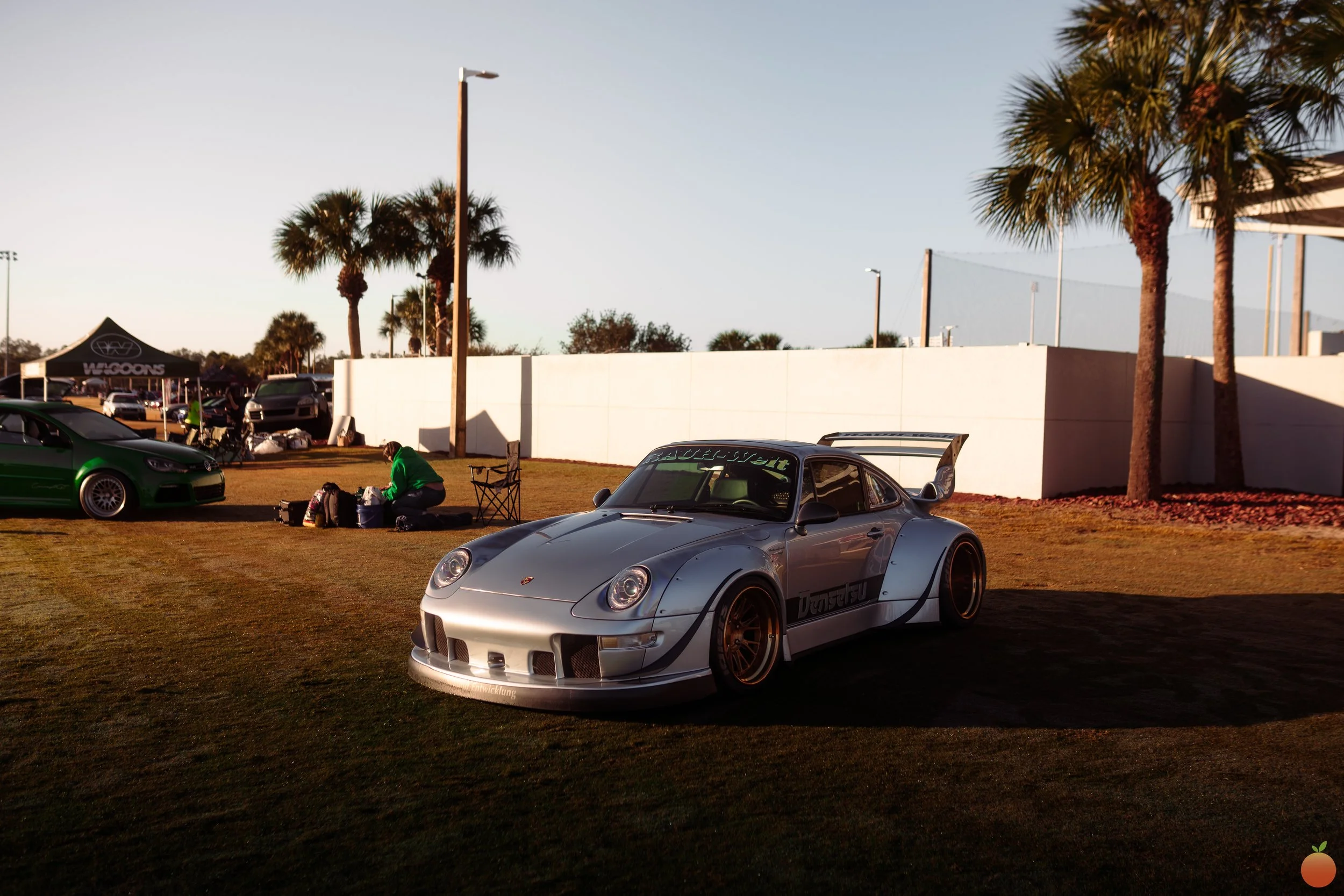 A silver sports car parked on grass at a car show, with palm trees, a white wall, a tent, and other cars in the background during sunset.