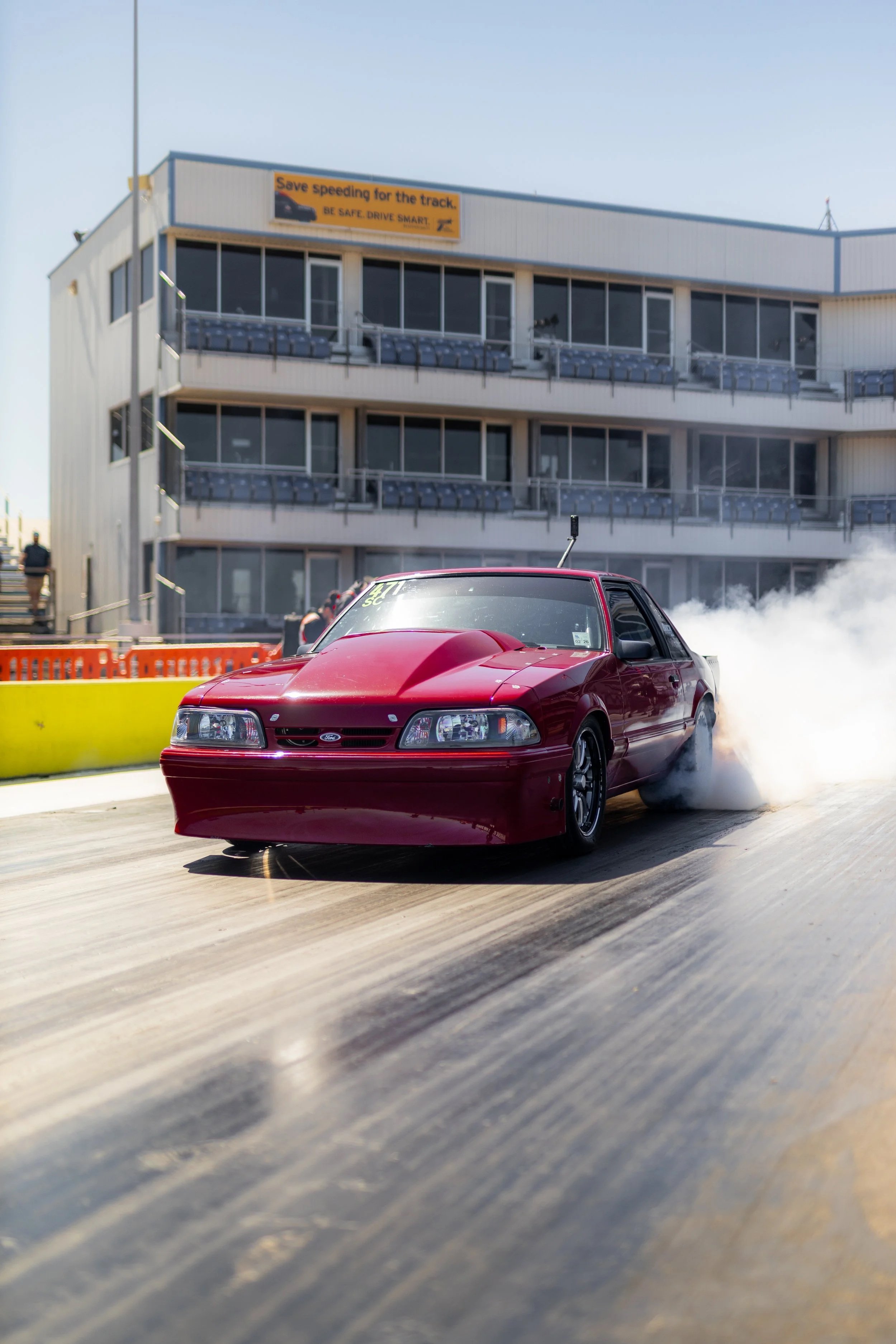 Red race car drifting on a race track with tires billowing smoke, a modern building with multiple levels and large windows in the background, and orange safety barriers along the track.