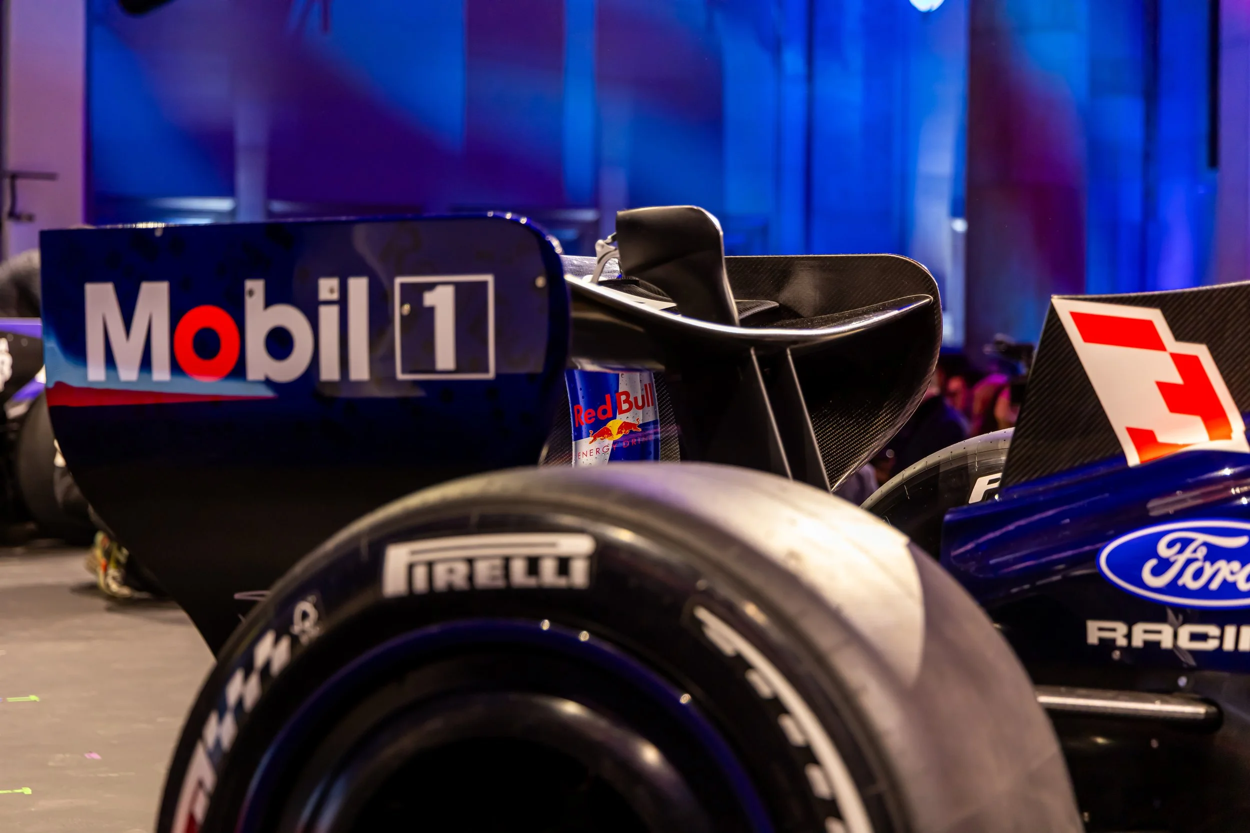 Close-up of a Formula 1 race car with sponsor logos including Mobil 1, Red Bull, Ford, and Pirelli, displayed indoors with a blue-lit background.