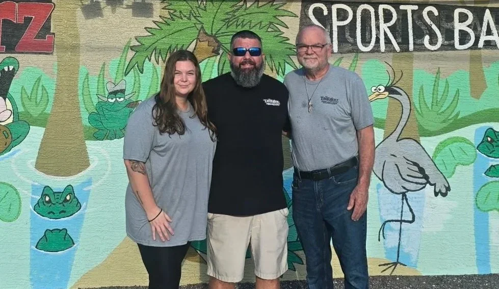 Three people standing in front of a mural with jungle animals and the words 'SPORTS BAR' in the background.