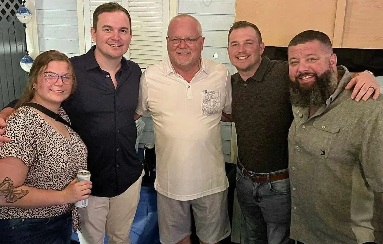 Group of five people smiling and standing together indoors, with a woman on the left and four men, including one in the center with glasses, all dressed casually.
