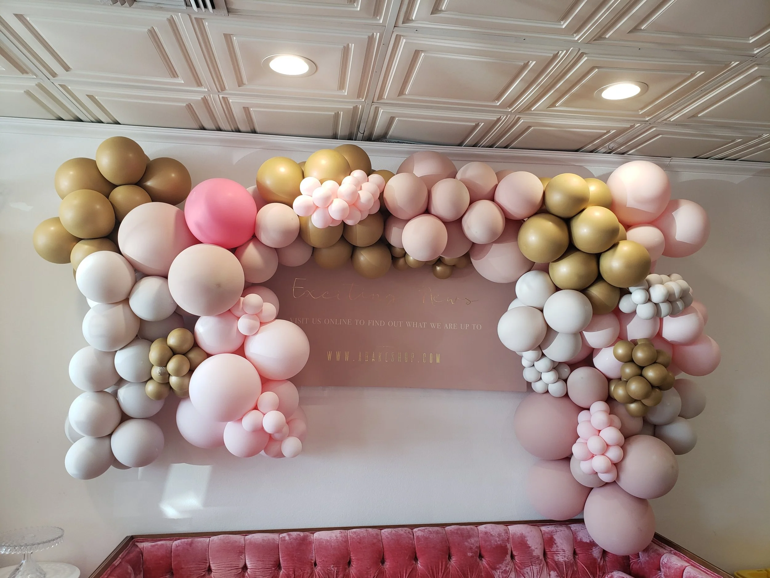 Balloon decoration with pink, white, and gold balloons arranged around a soft pink backdrop and a pink velvet sofa.