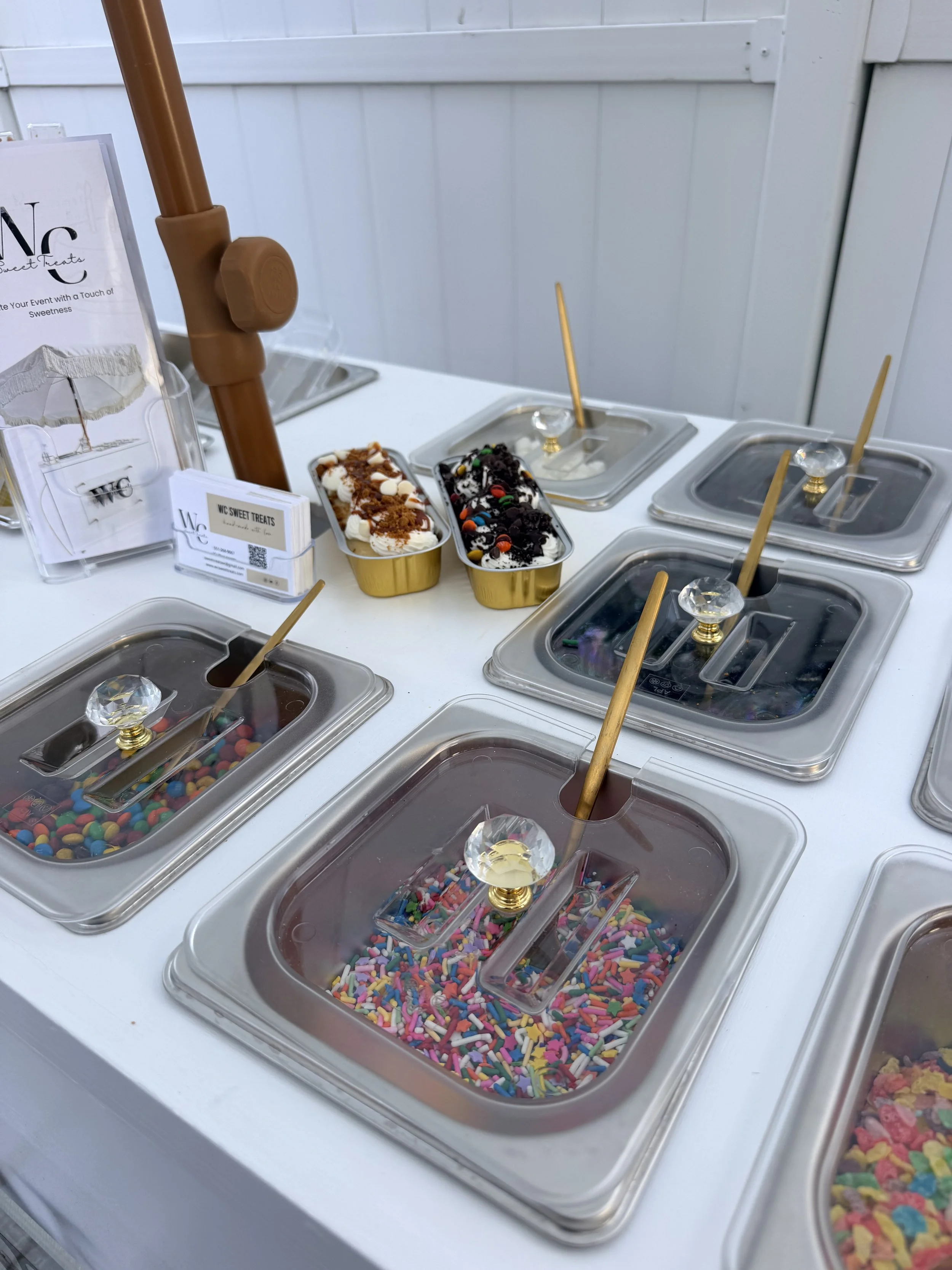 A dessert display with various toppings and candies, including sprinkles, chocolate chips, and candies, in separate containers, with ice cream in cones and business cards on the side.