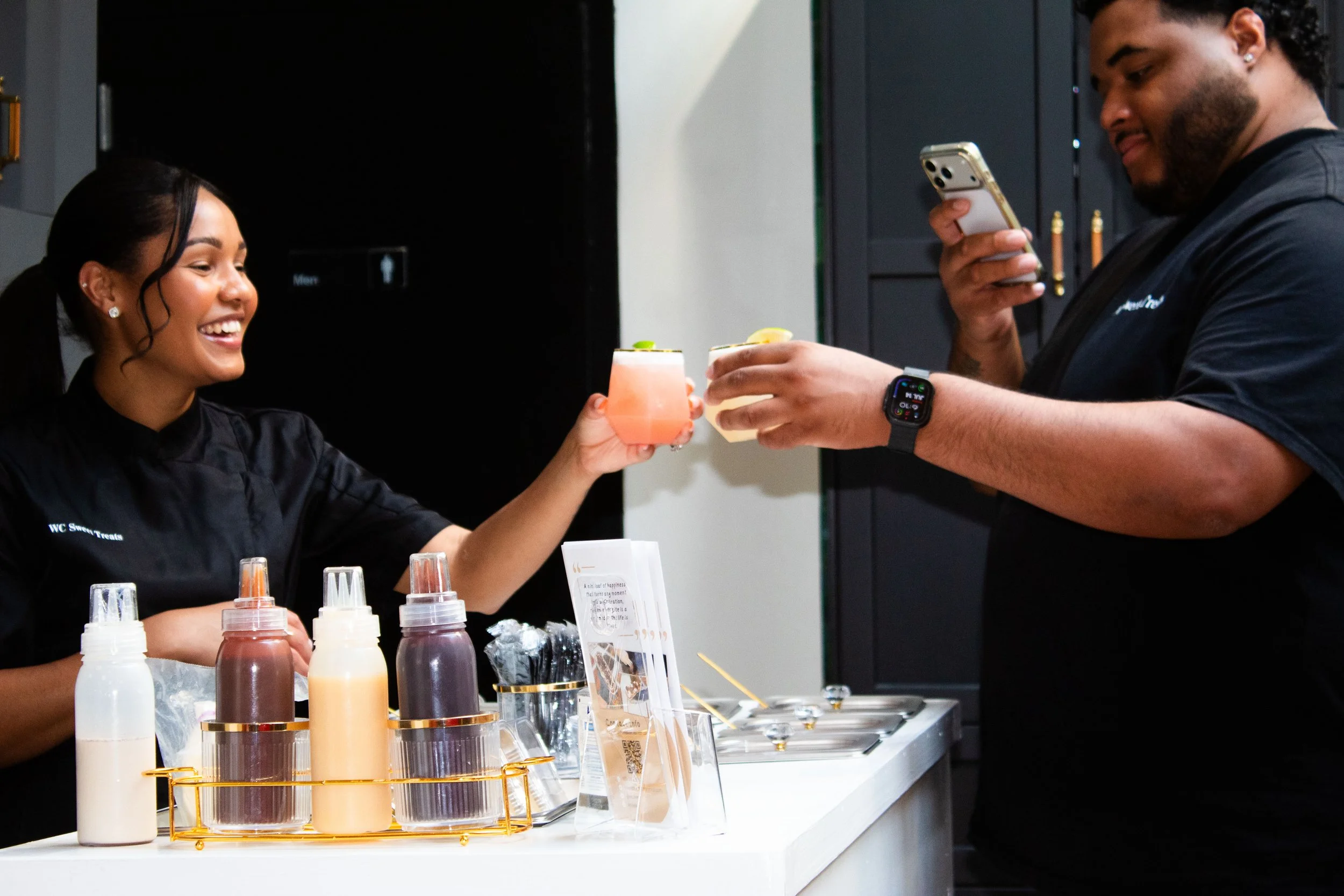 A woman with dark hair, earrings, and a black shirt smiling as she hands a pink cocktail to a man with dark hair, a beard, and earrings, who is looking at his phone. They are at a counter with bottles of various syrups and a sign, in a modern indoor setting.