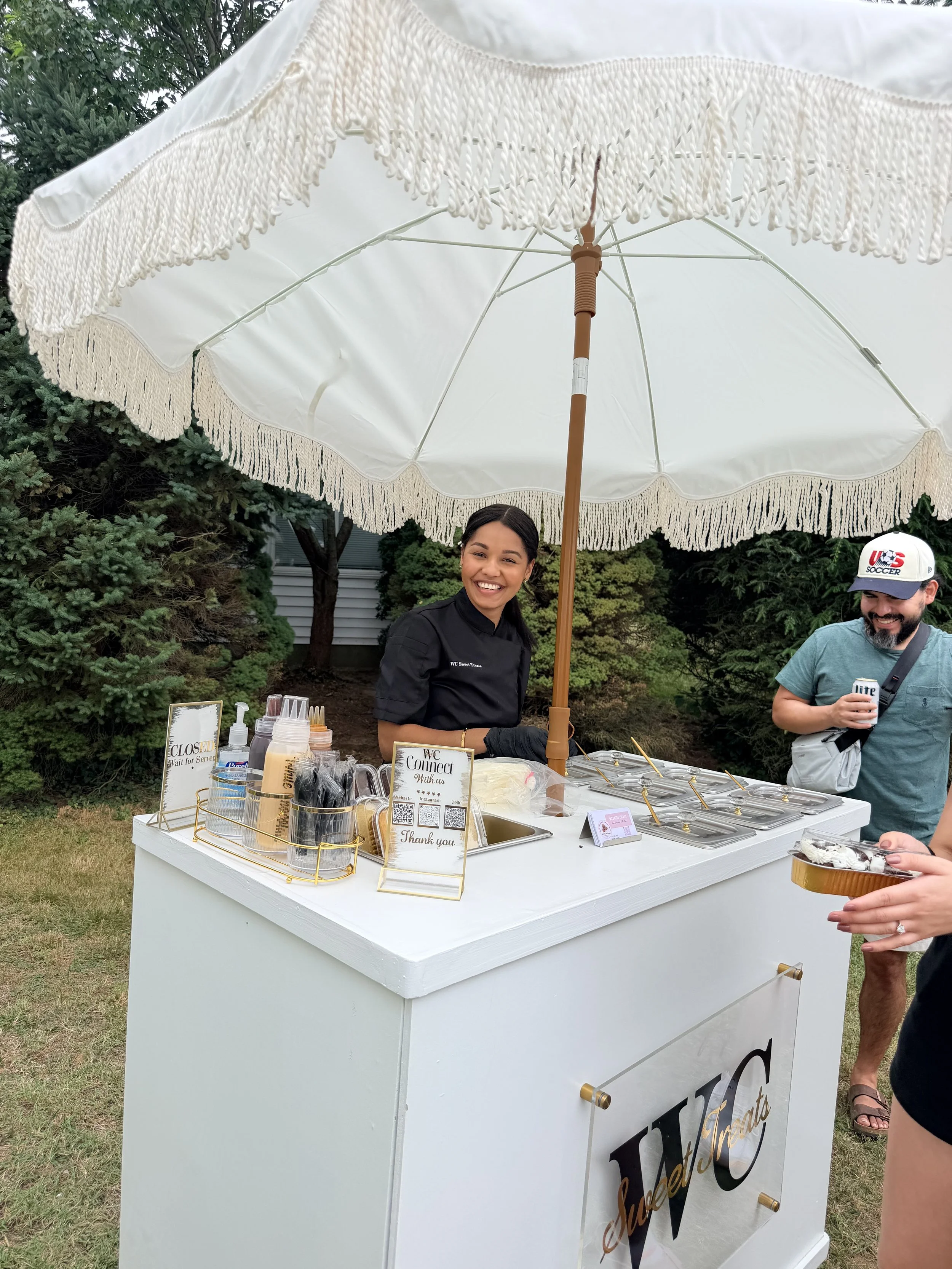 A women working at a white dessert stand under a white umbrella with fringes. There are various dessert items and signs on the stand. A man is seen beside the stand holding a cup.