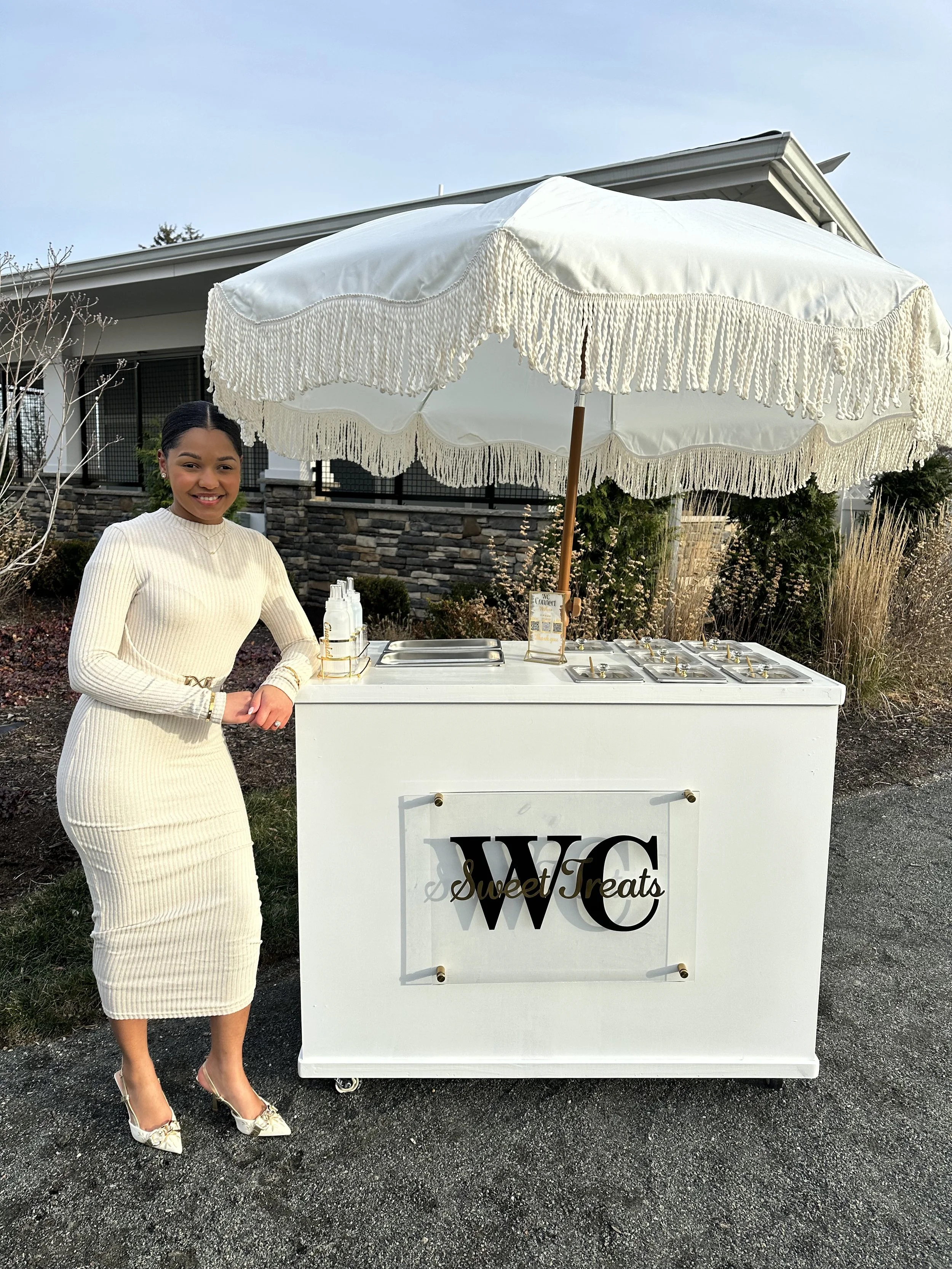 A young woman stands next to a white sweets cart under a large white parasol with fringe. The cart has a sign with the logo "WC" and the words "Sweet Treats." She is smiling, wearing a cream-colored dress and matching high heels, outdoors in front of a house with stone accents.